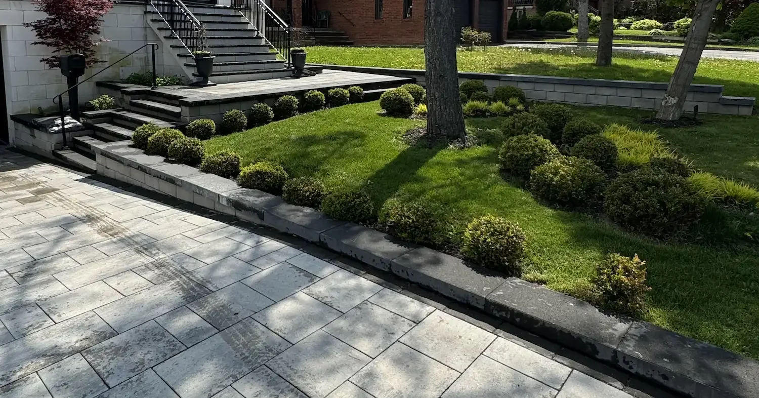 A landscaped front yard with stairs leading to a house, bordered by neatly trimmed bushes and a grassy area with trees, and a stone patio in the foreground.