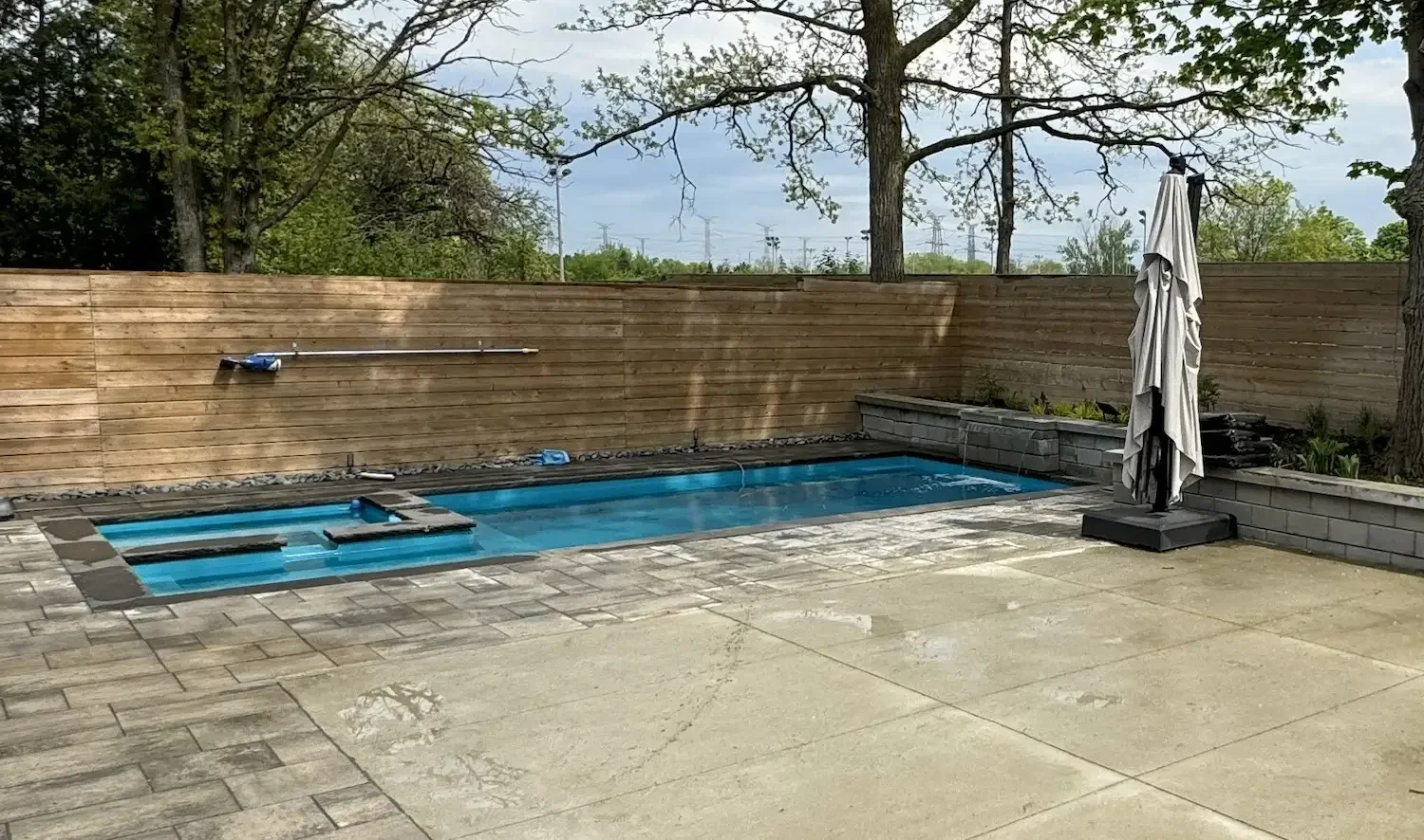 A small backyard patio with a rectangular swimming pool, a wooden privacy fence, a closed patio umbrella wrapped in fabric, and some trees with green leaves in the background.