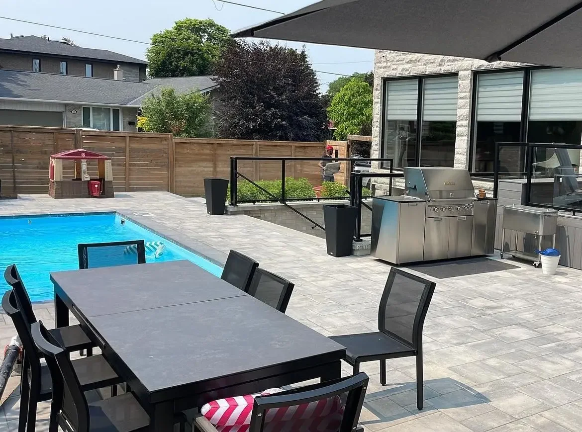 Backyard patio with a swimming pool, outdoor dining table with black chairs, a stainless steel grill, and a small playhouse with a red umbrella in the background. Fence surrounds the yard, and there are houses with gray roofs nearby.