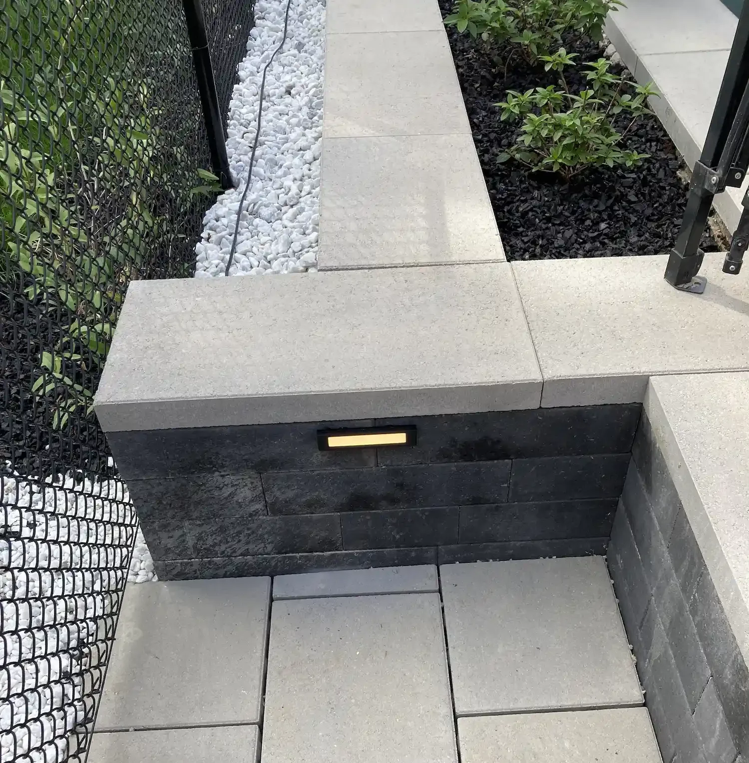 Elevated outdoor walkway with large beige tiles, a stone wall with a built-in light, a garden with plants, and a black metal fence.