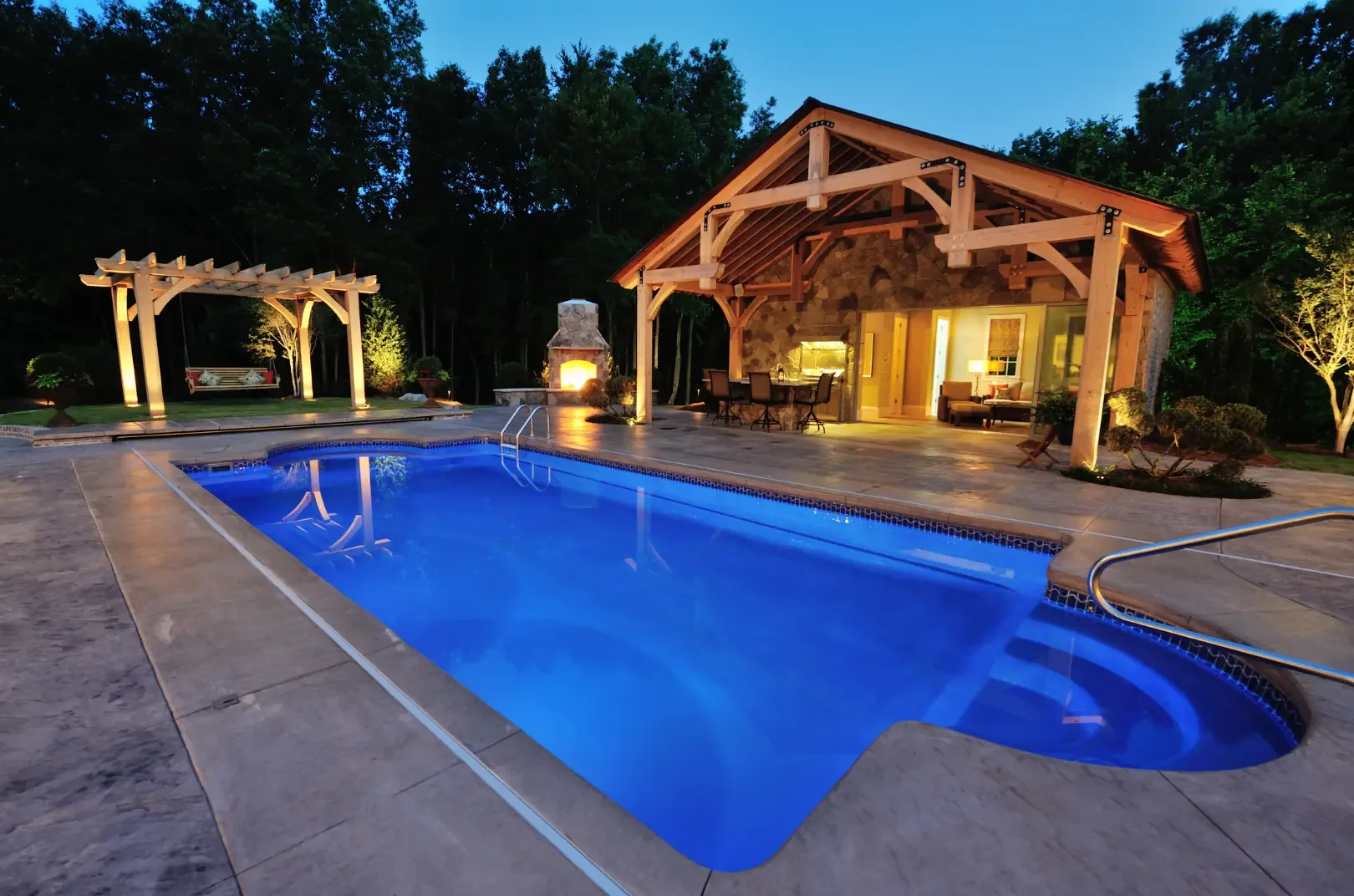 Luxury backyard with illuminated swimming pool, stone fireplace, outdoor seating, and a wooden pavilion in a wooded area at dusk.