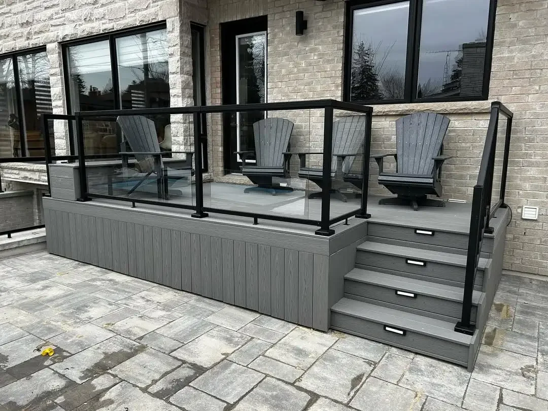 Gray wooden deck with three steps leading up to it, black metal and glass railing, four Adirondack chairs, brick house background, stone patio in foreground.