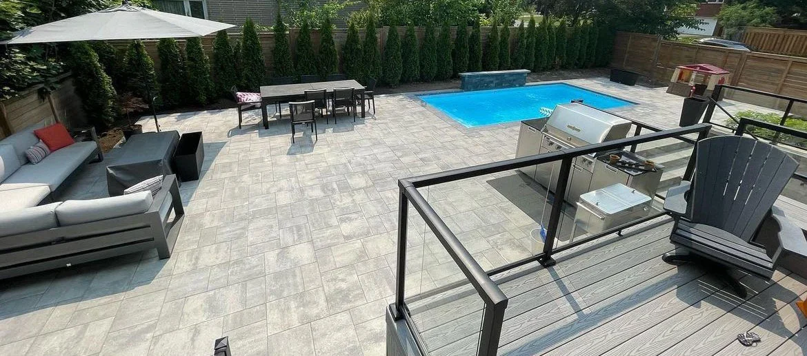 Backyard with a swimming pool, outdoor seating area with a lounge sofa, dining table with chairs, and a BBQ grill, enclosed by a wooden fence and surrounded by tall evergreen trees.