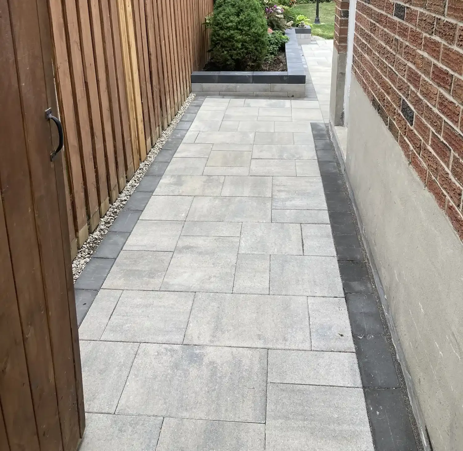 A paved walkway with light gray rectangular tiles bordered by a darker gray strip, flanked by a wooden fence and a brick wall, with a small raised garden bed at the end containing plants and flowers.