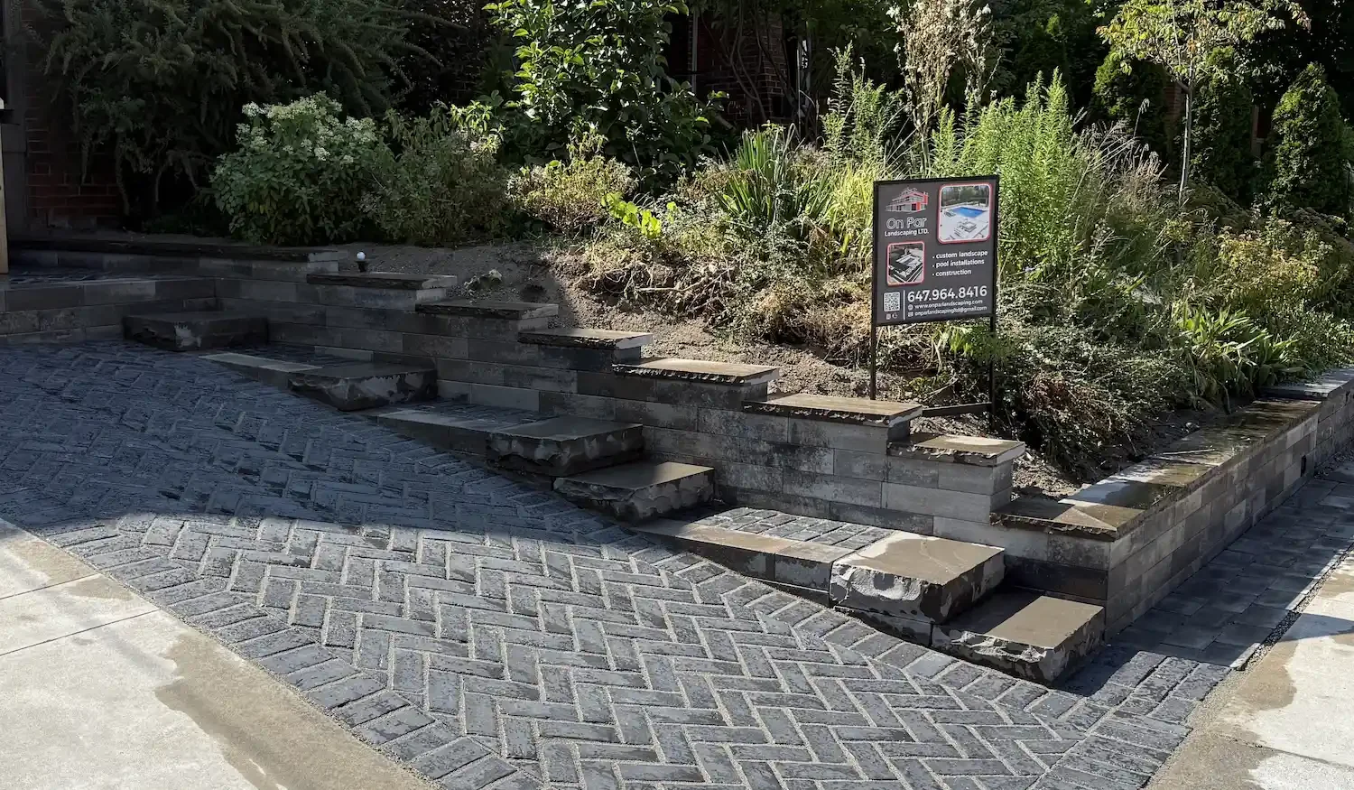 A landscaped outdoor space with a brick retaining wall, stone steps leading up to the garden, and a sign advertising landscaping services. The area features plants and a small water feature on the right.