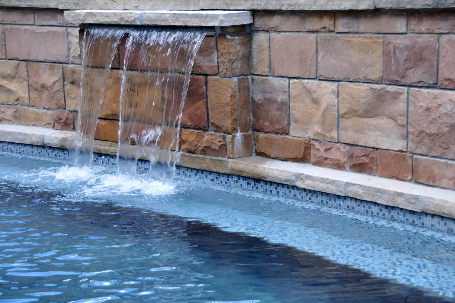 Water flowing from a small waterfall in a swimming pool with a stone and brick wall background.