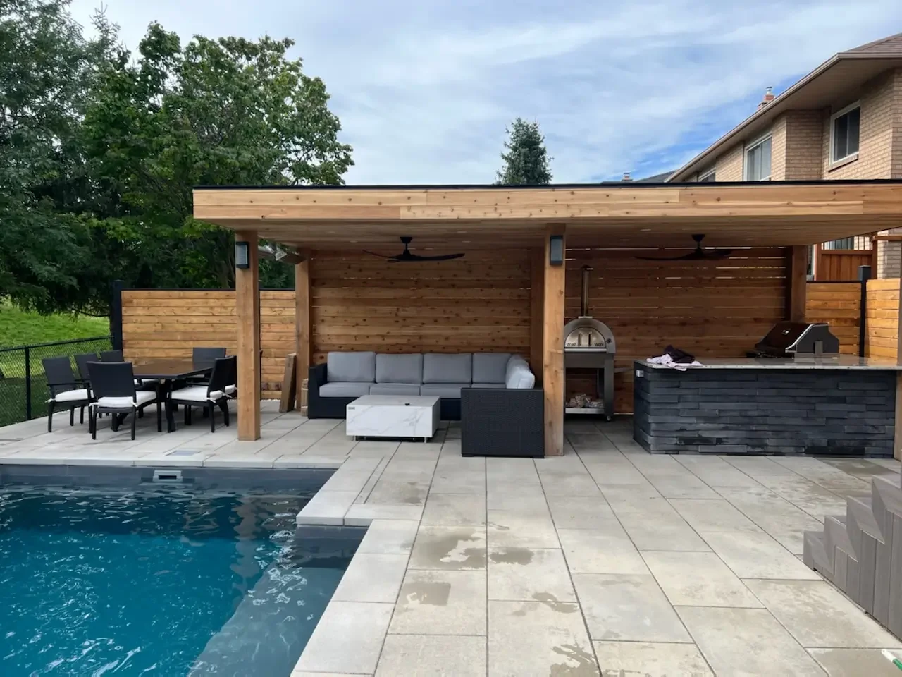 Outdoor backyard area with a swimming pool, black dining table with chairs, gray sofa, white and black marble coffee table, pizza oven, outdoor kitchen with a black chimney, and a wooden privacy fence, with a house and trees in the background.