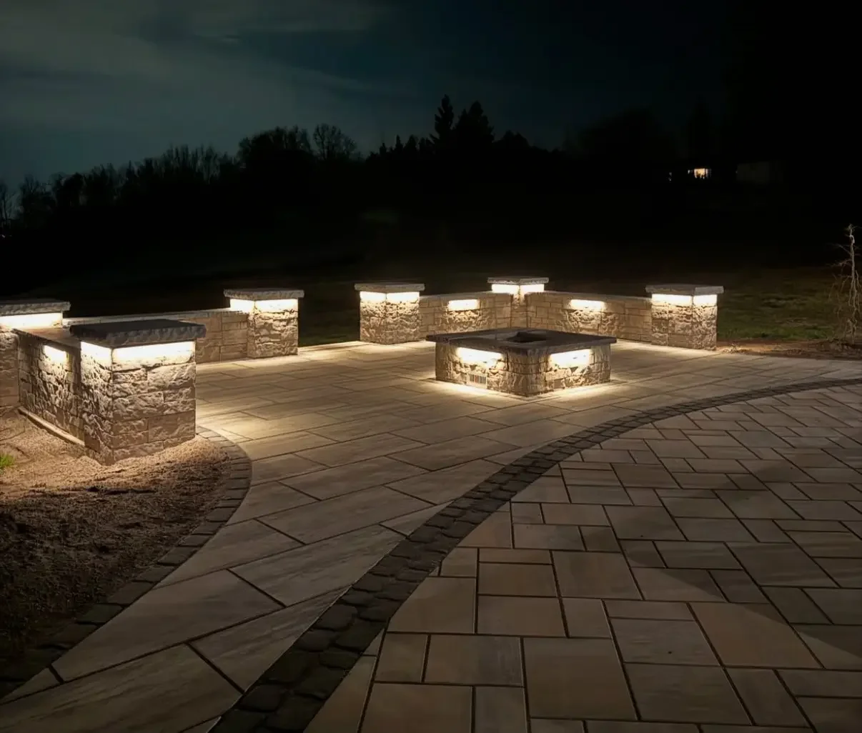 Nighttime view of a decorated outdoor patio with stone walls and benches illuminated by built-in lighting, surrounded by a brick-patterned walkway and dark landscape.