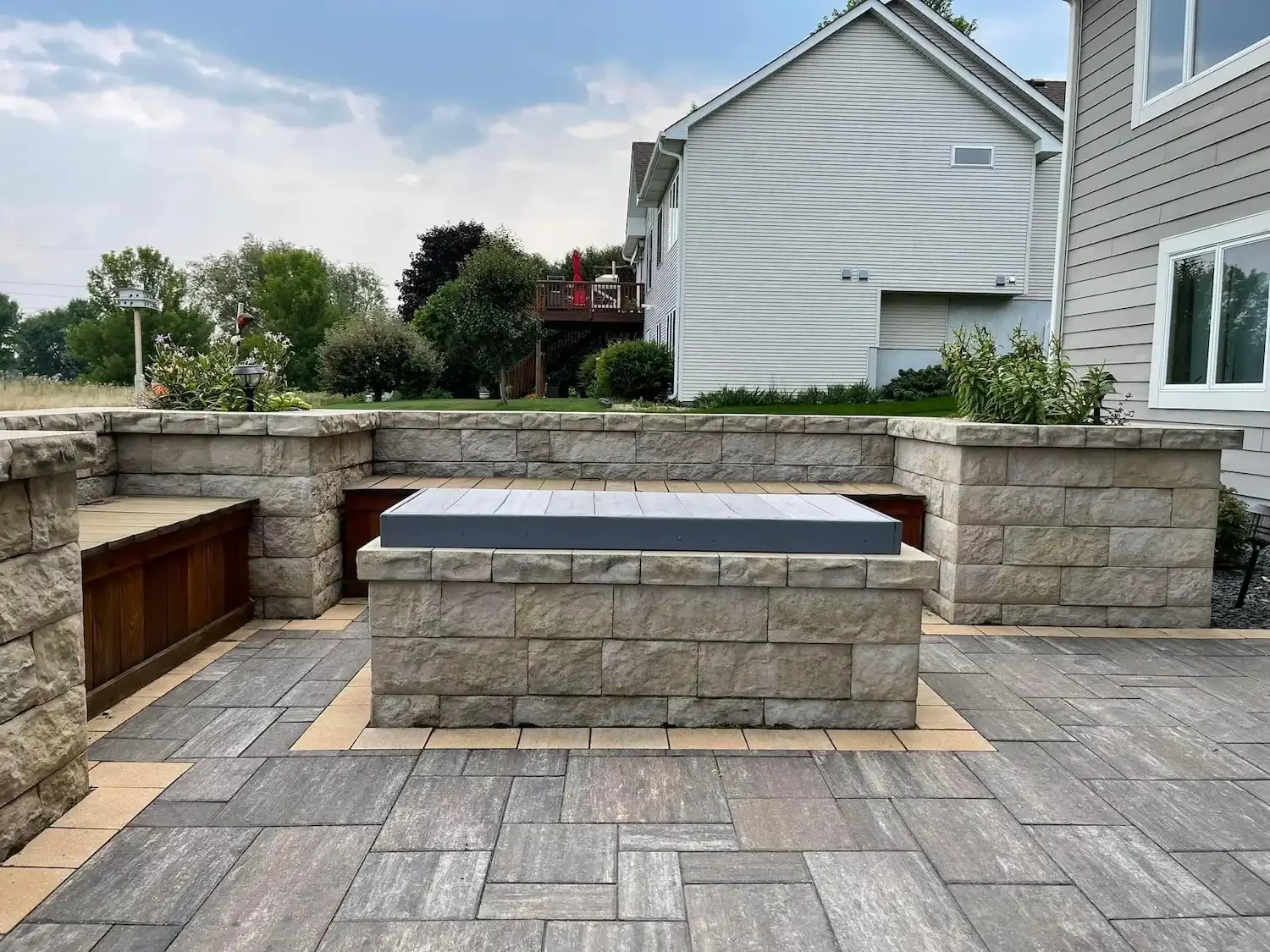 Backyard patio with stone and wood seating walls, a tiled floor, and a raised wood fire pit in the center. There are houses with siding and a deck with a red umbrella in the background, along with trees and shrubs under a partly cloudy sky.