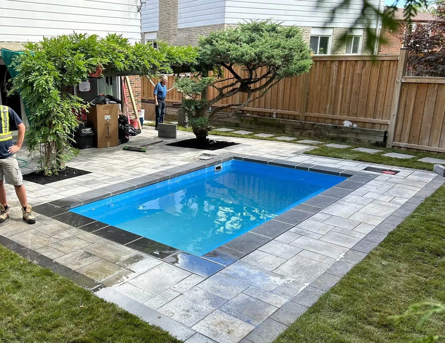 A backyard with a small rectangular swimming pool, surrounded by stone pavers and a grassy area. There are two people working near a shed, a young man in the foreground, and an older man in the background. A small leafy tree is growing along the fenc