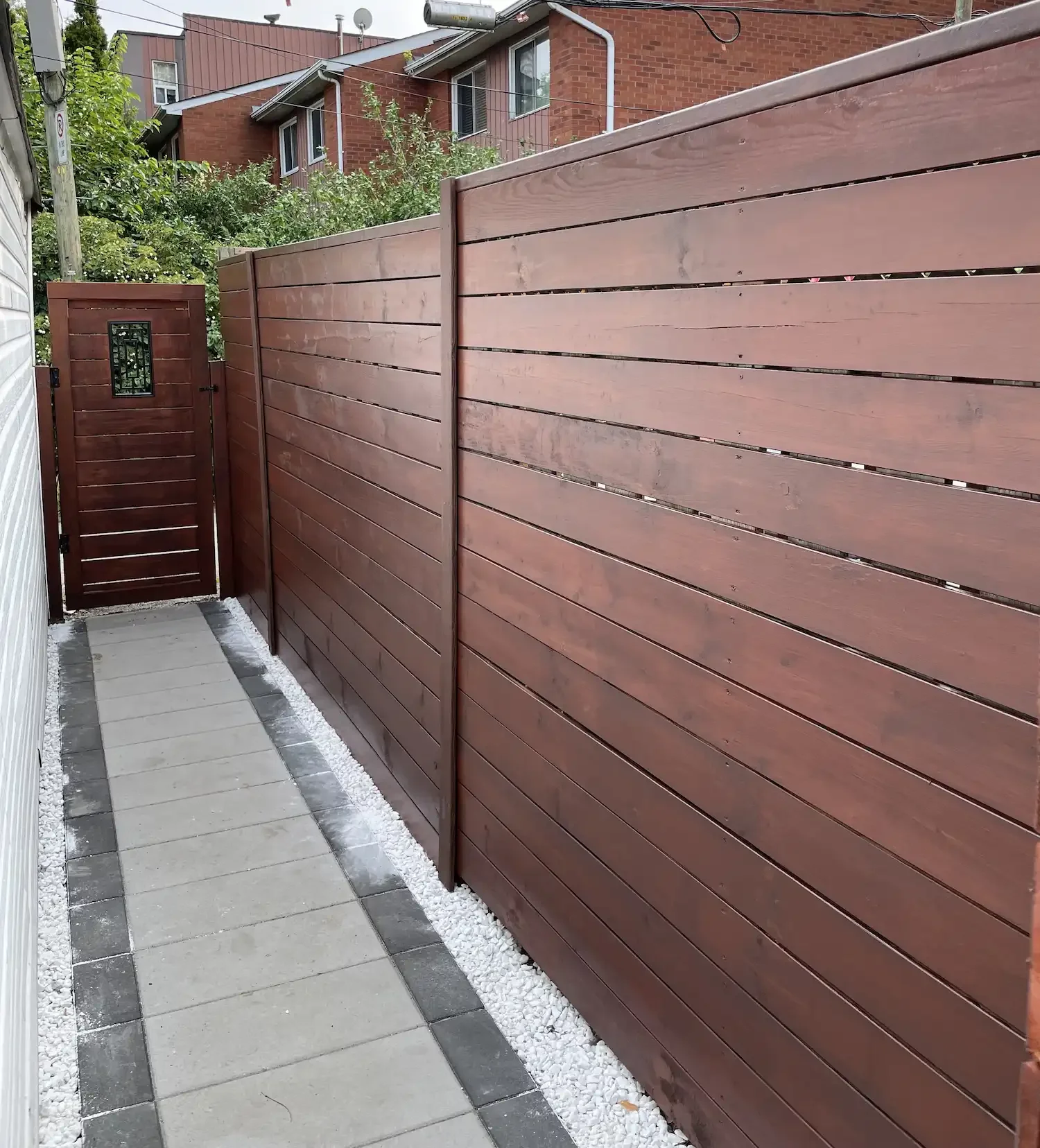 A brown wooden privacy fence along a concrete walkway with white gravel at the base, topped with a matching gate, with houses and greenery in the background.