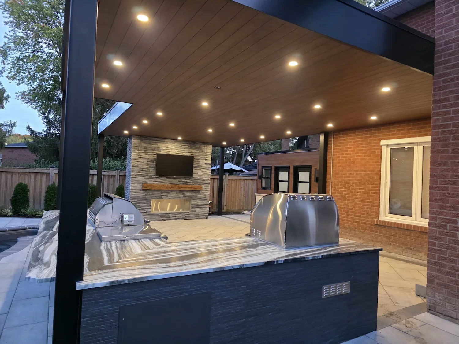 Outdoor kitchen with granite countertop, built-in grill, and stainless steel appliances, under a wooden ceiling with recessed lighting. There is a stone fireplace, a wall-mounted TV, and a fenced backyard with trees and landscaping.