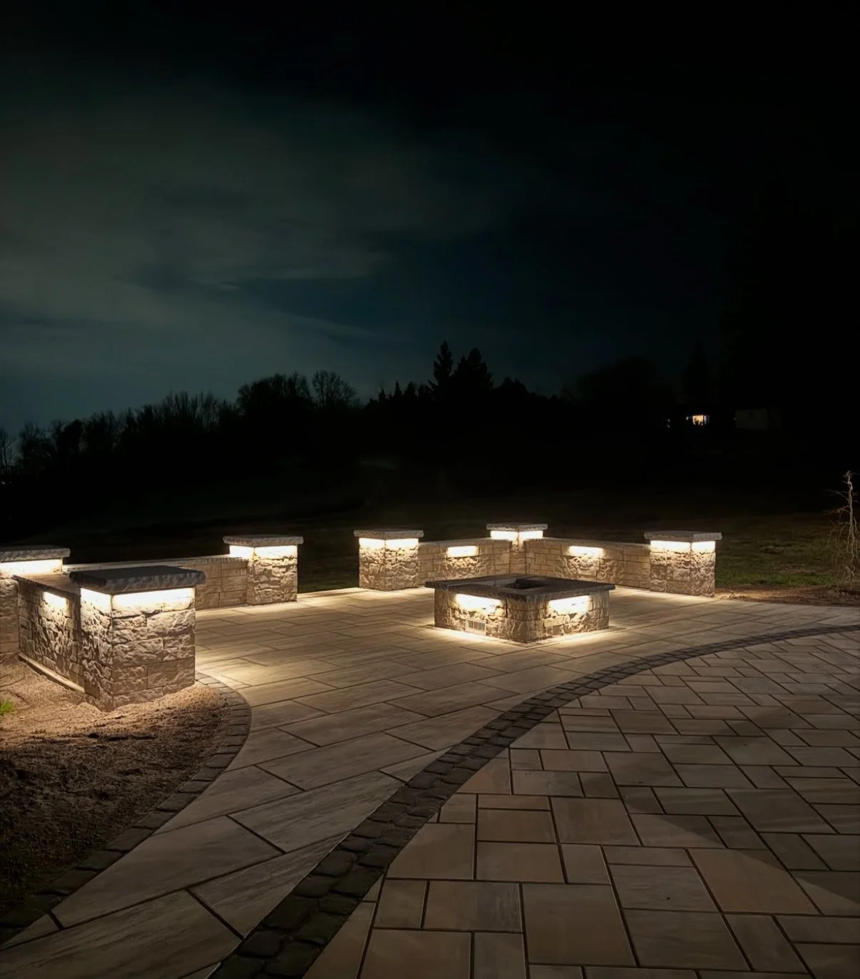 Outdoor stone patio with illuminated stone pillars and firepit at night.