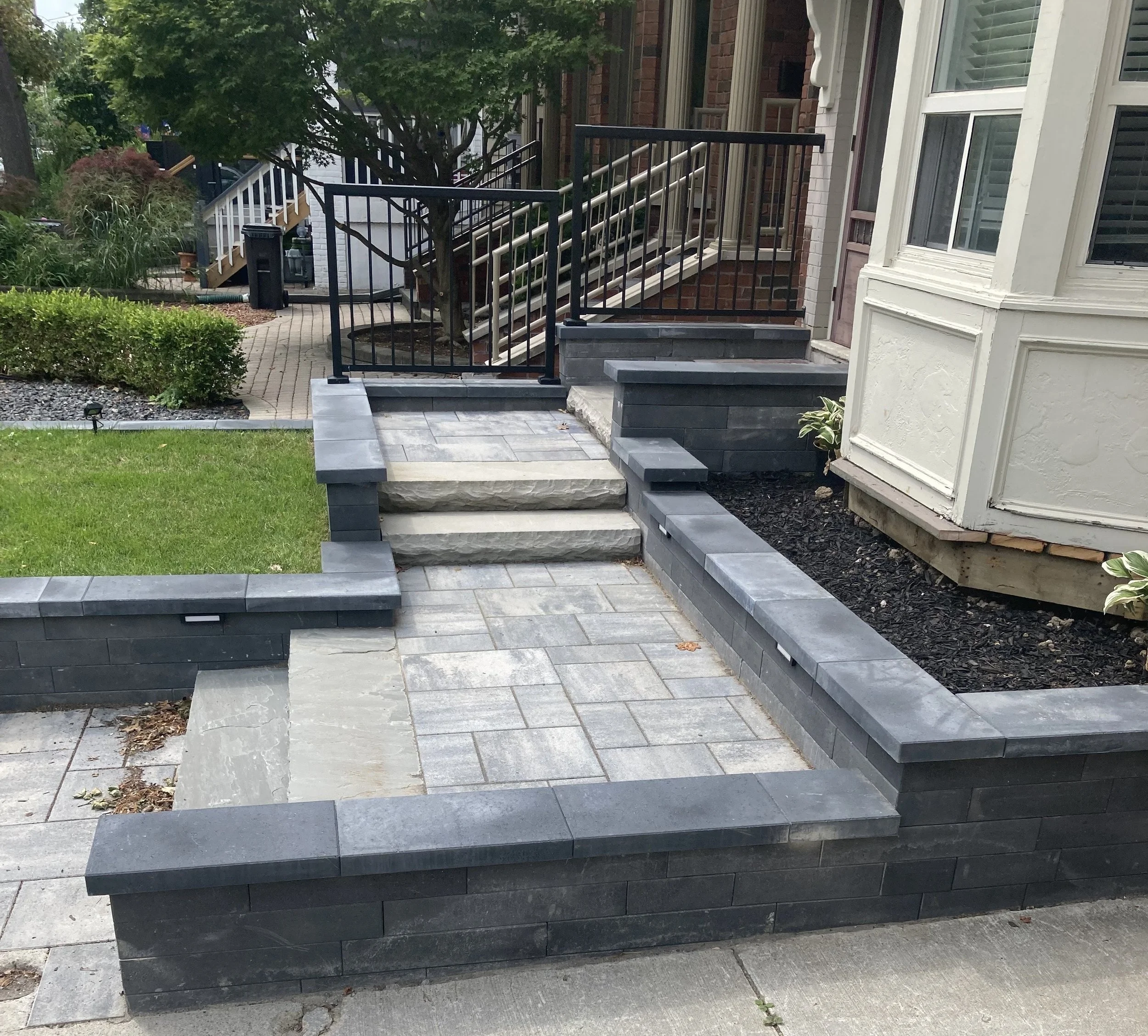 Residential front porch with stairs, black metal railing, gray stone and concrete pavement, landscaped garden, and neighboring brick and wood houses.