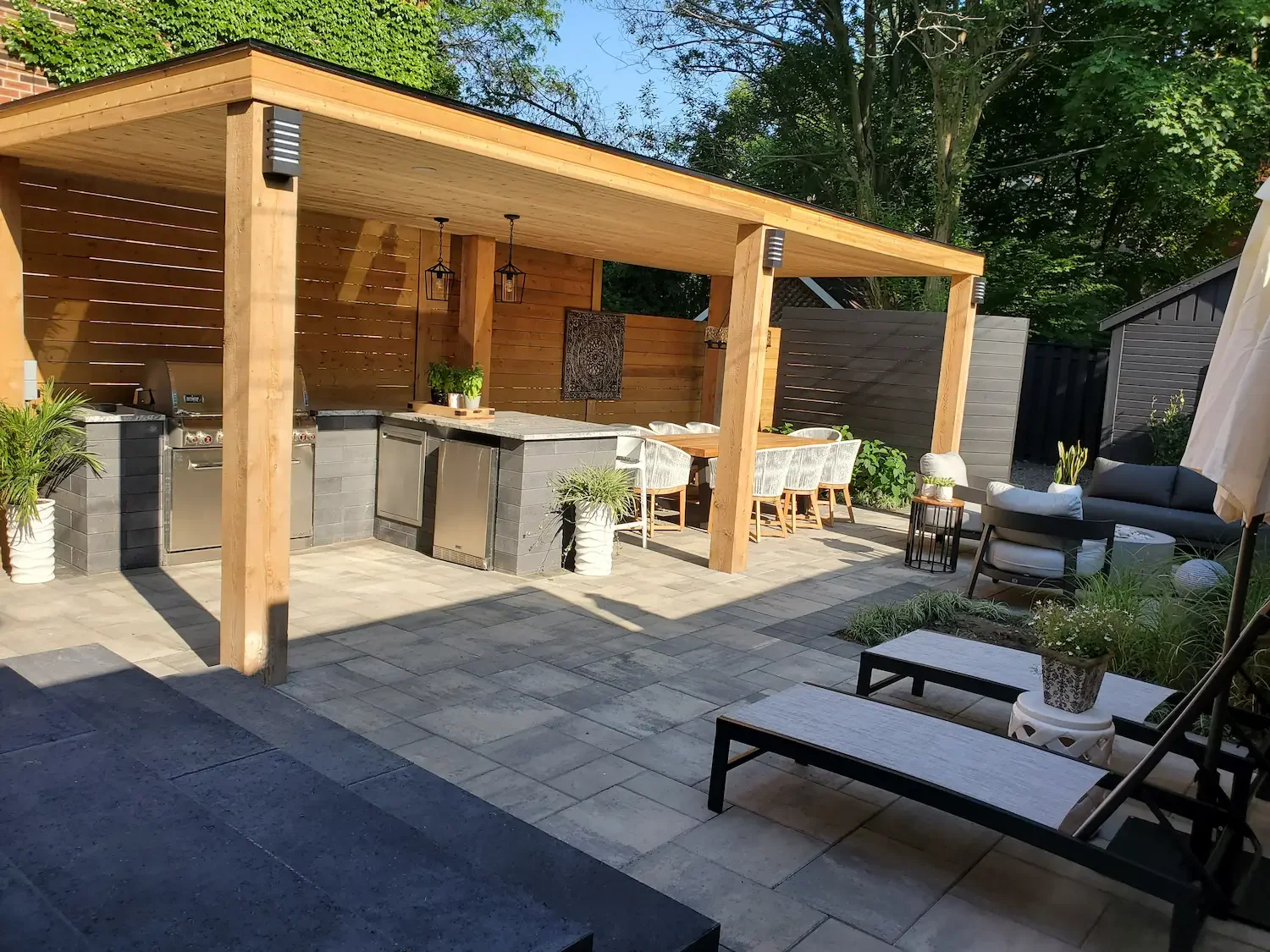 A modern outdoor patio with a covered cooking and dining area, featuring a built-in countertop, grill, potted plants, a dining table with chairs, and a seating area with lounge chairs, all with greenery and trees in the background.