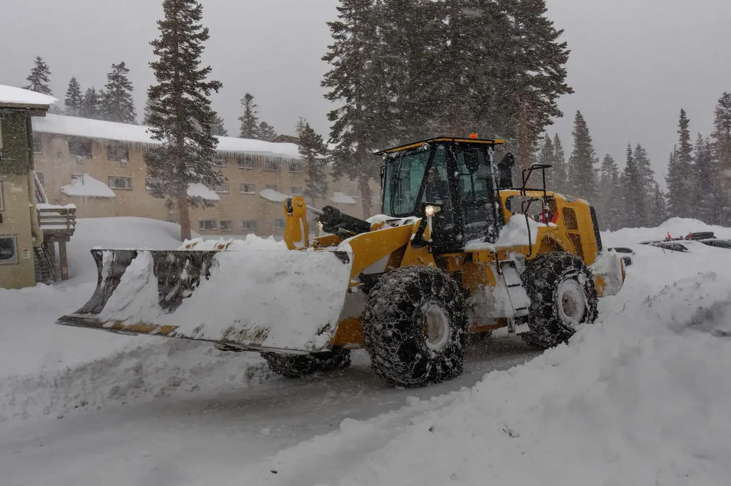A yellow snowplow clearing snow in a snowy environment with trees and building in the background.