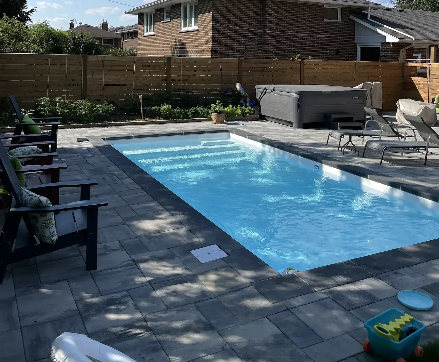 Backyard with a swimming pool, lounge chairs, hot tub, and a wooden fence, with houses in the background.