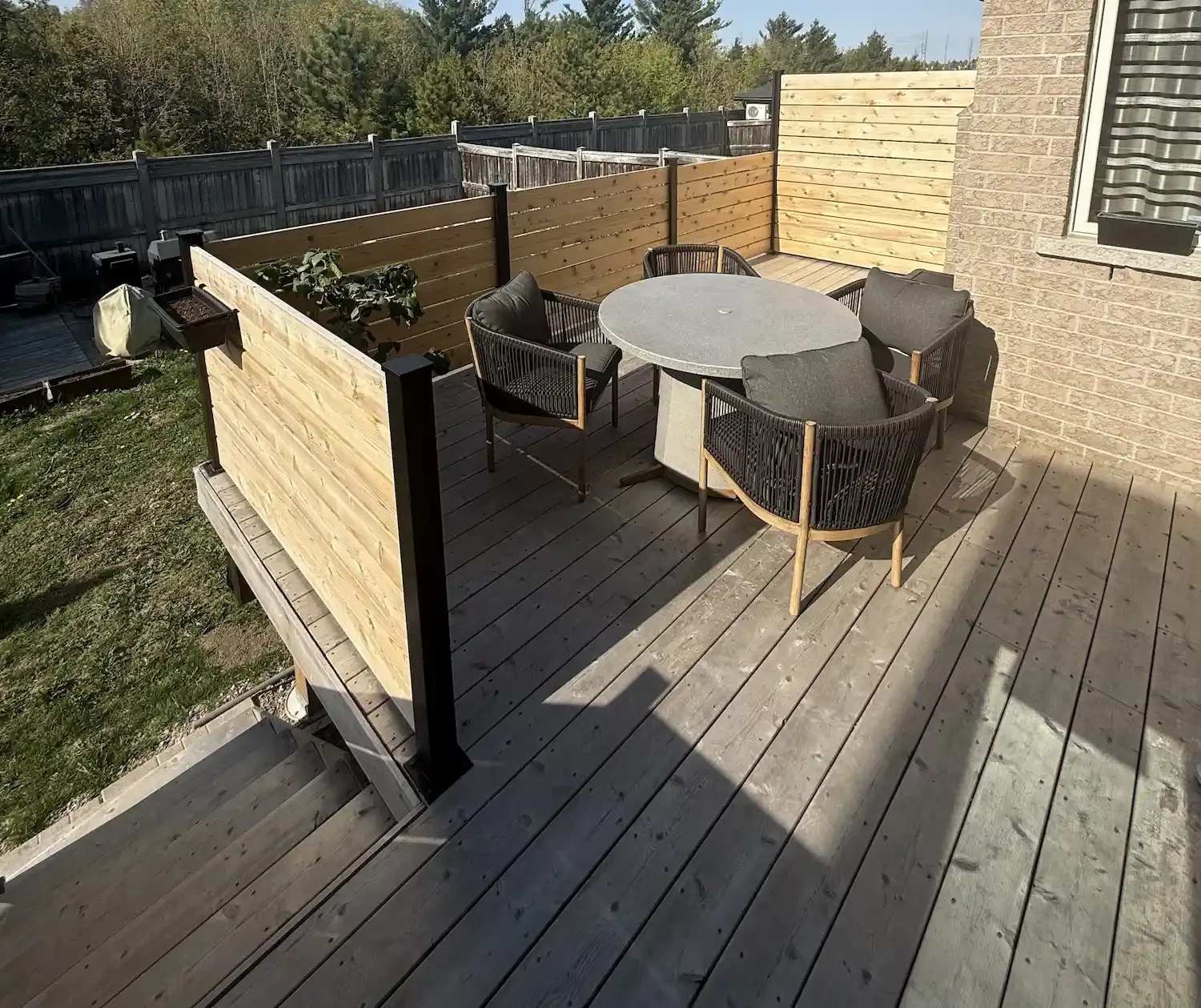 Wooden backyard deck with round table and four chairs, surrounded by a wooden privacy fence, attached to a brick house, on a sunny day.
