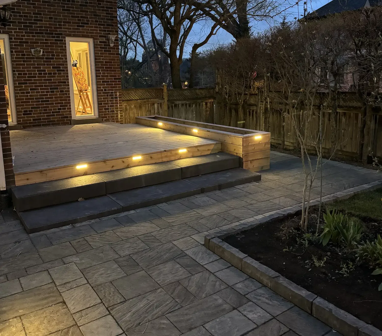 A backyard patio area at dusk with a newly built wooden deck and stairs, lit by embedded ground lights, surrounded by a stone-paved area, a fenced backyard, a small garden bed with plants, and a brick house with a door and windows.
