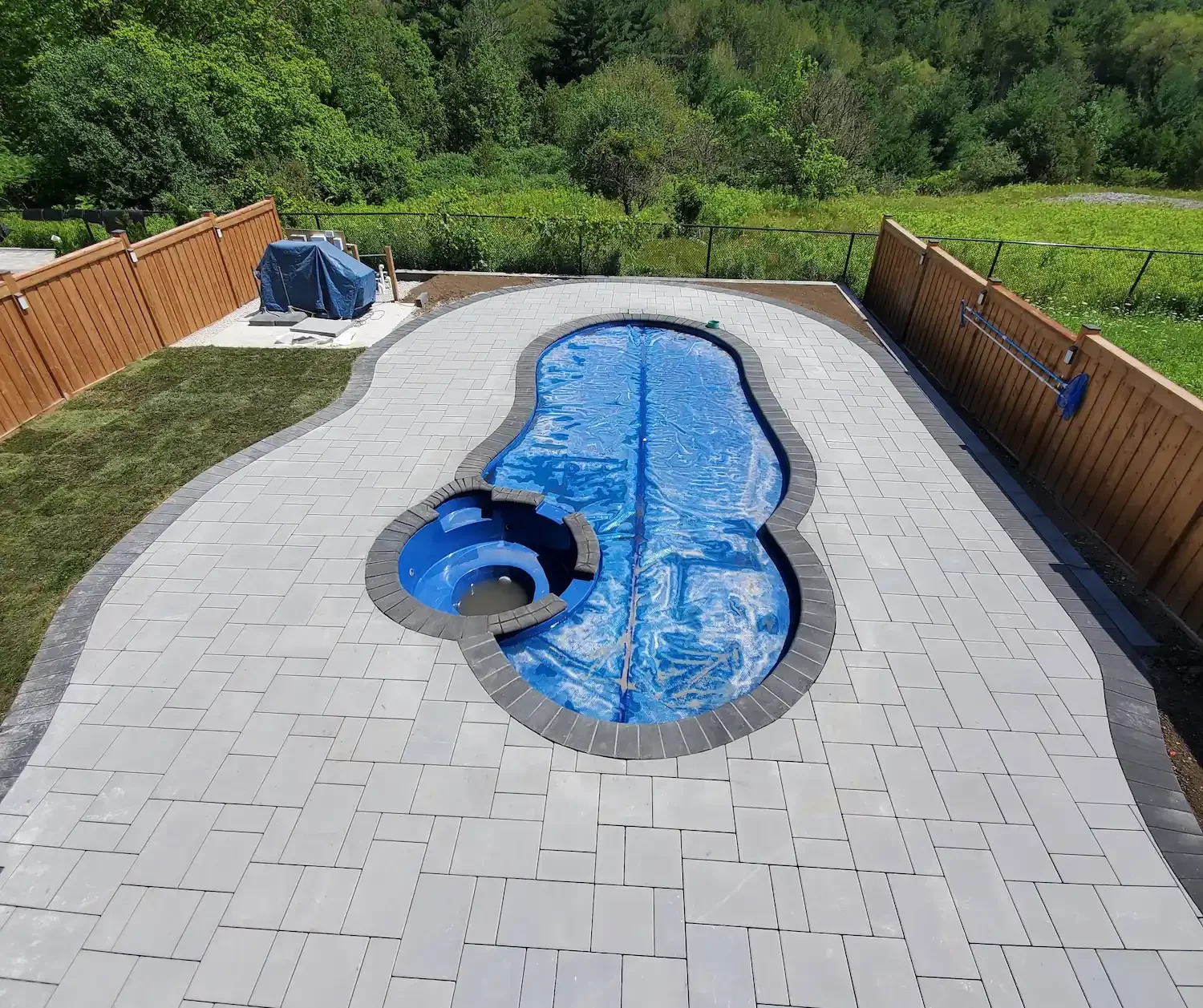 Above view of a backyard with a fenced yard, a covered hot tub, and a swimming pool with a slide, surrounded by gray pavers and a patch of grass in the corner, and greenery beyond the fence.