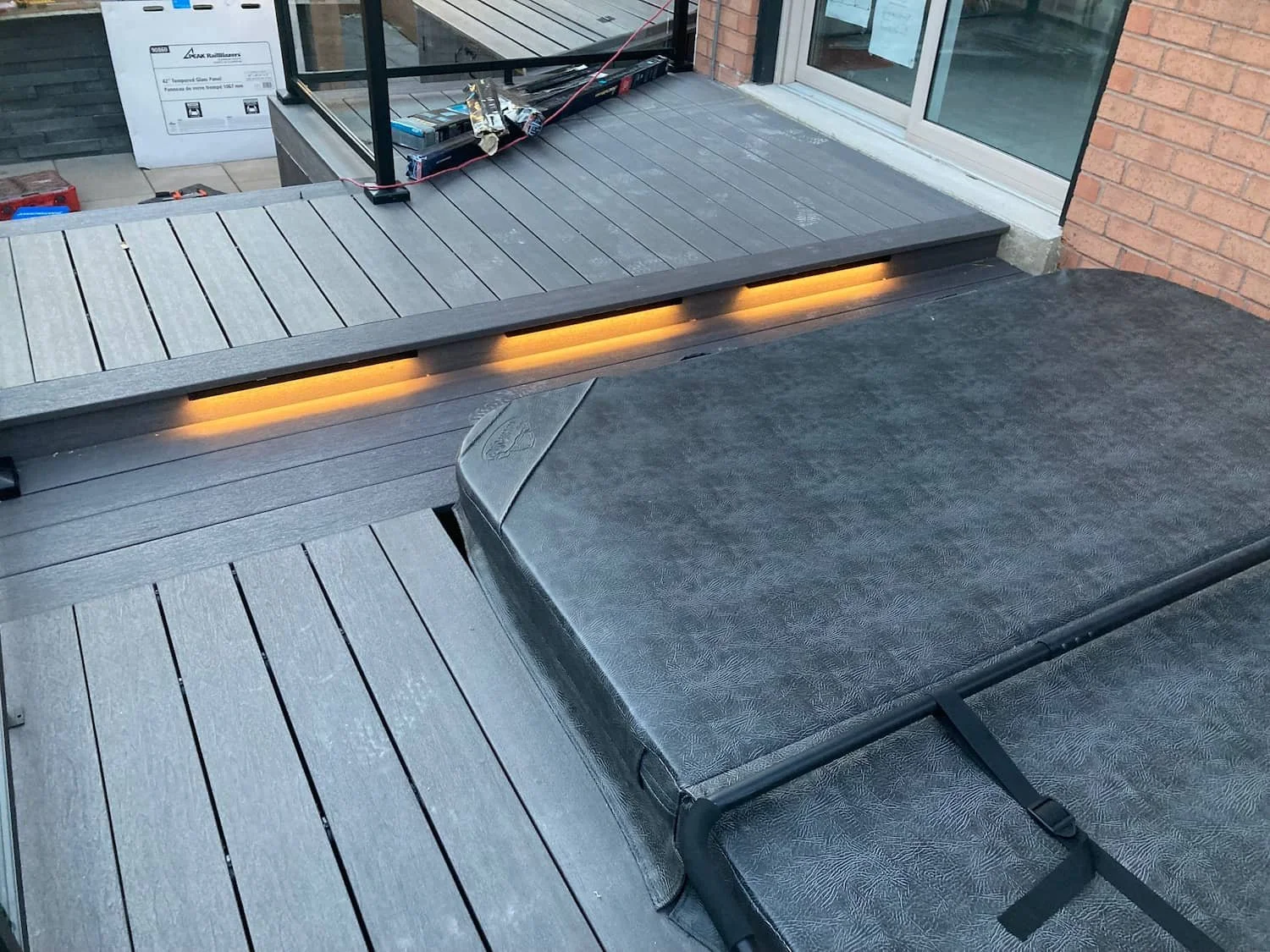 An outdoor deck area with wooden flooring, a skateboard with a textured black surface, a glass railing, a sliding door, and construction materials in the background.