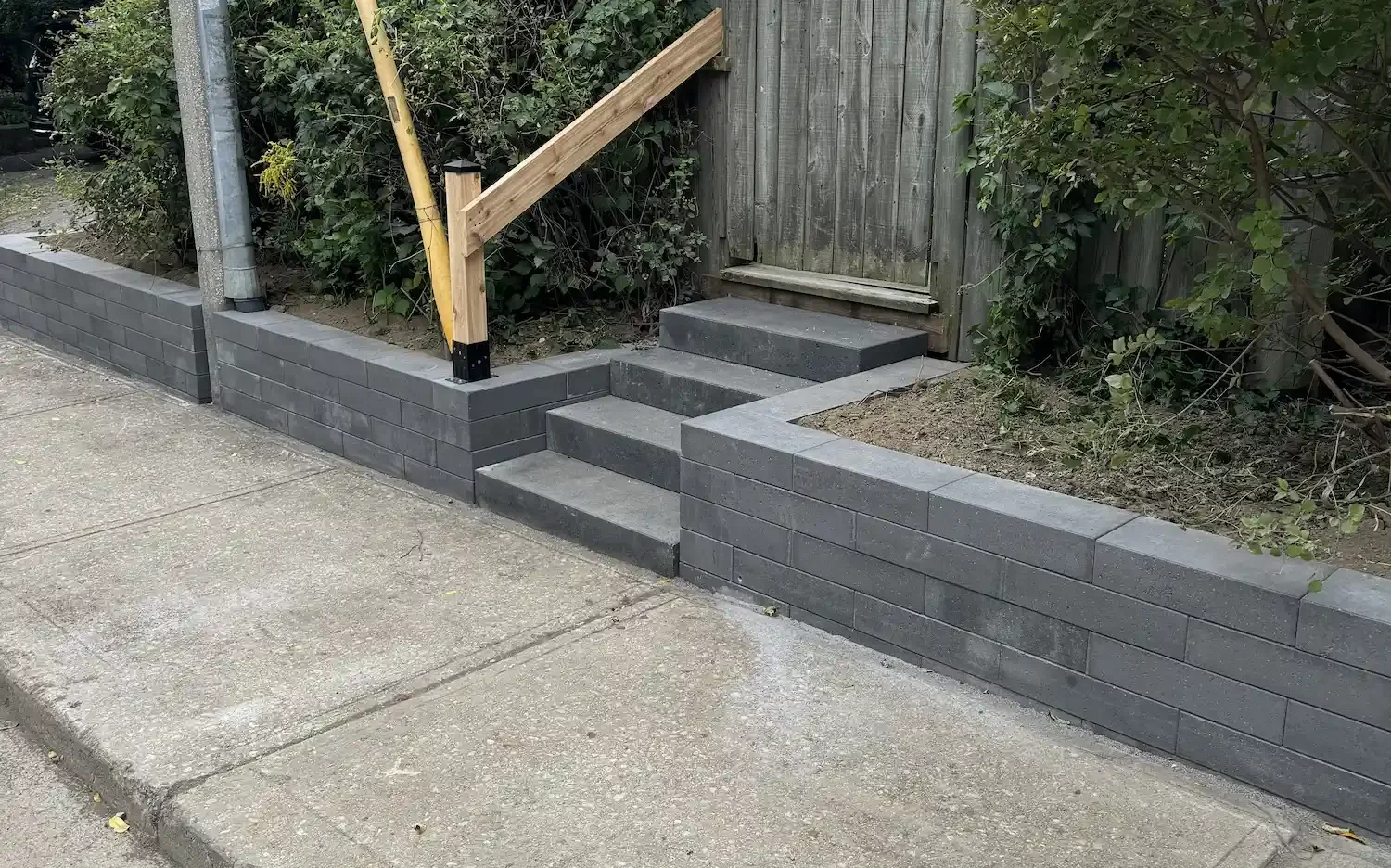 Newly built gray brick retaining wall with steps, adjacent to sidewalk, with wooden fencing and plants behind.