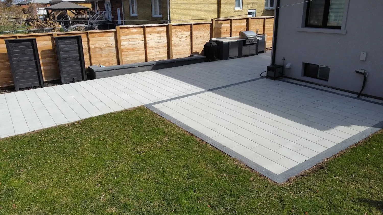 Backyard patio area with white tiles, a small grassy section, and a wooden privacy fence. There is a grill and some equipment against the house.