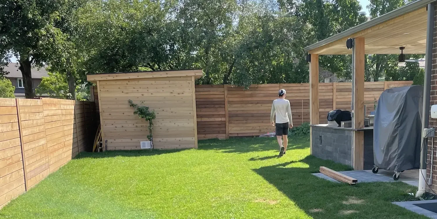 A backyard with green grass, a wooden shed, a person walking away wearing a cap, long sleeve shirt, shorts, and sneakers, a covered outdoor kitchen area with a grill, and a wooden fence.