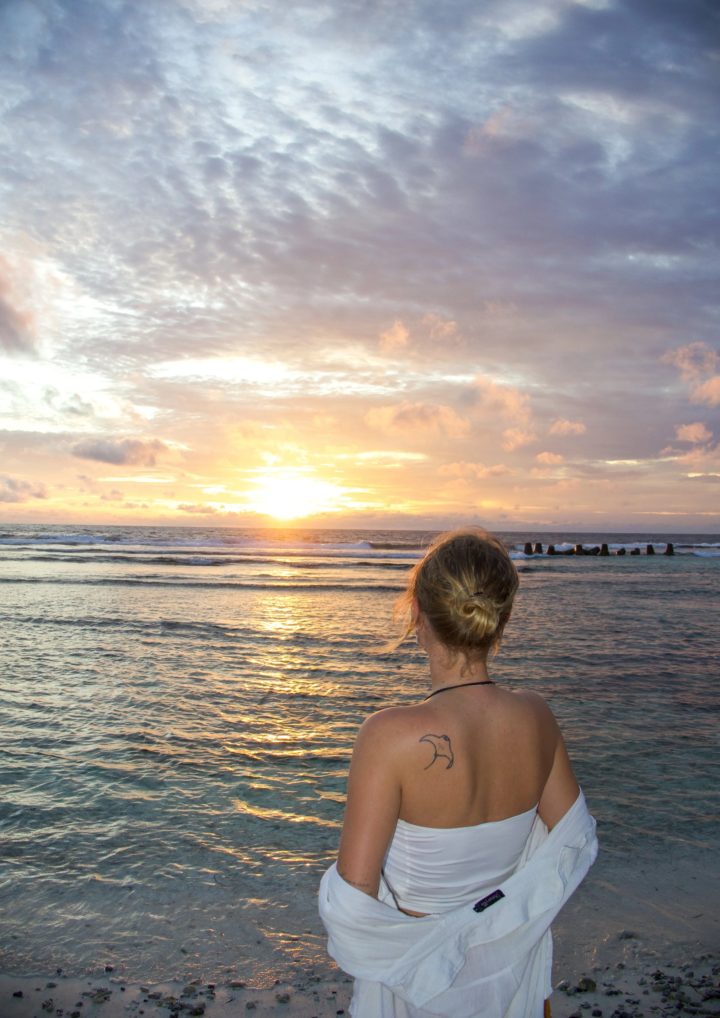 A woman with blonde hair tied in a bun standing on the beach, looking at the sunset over the ocean, wearing a white strapless top and a white shirt draped around her shoulders, with a moon tattoo on her upper back.