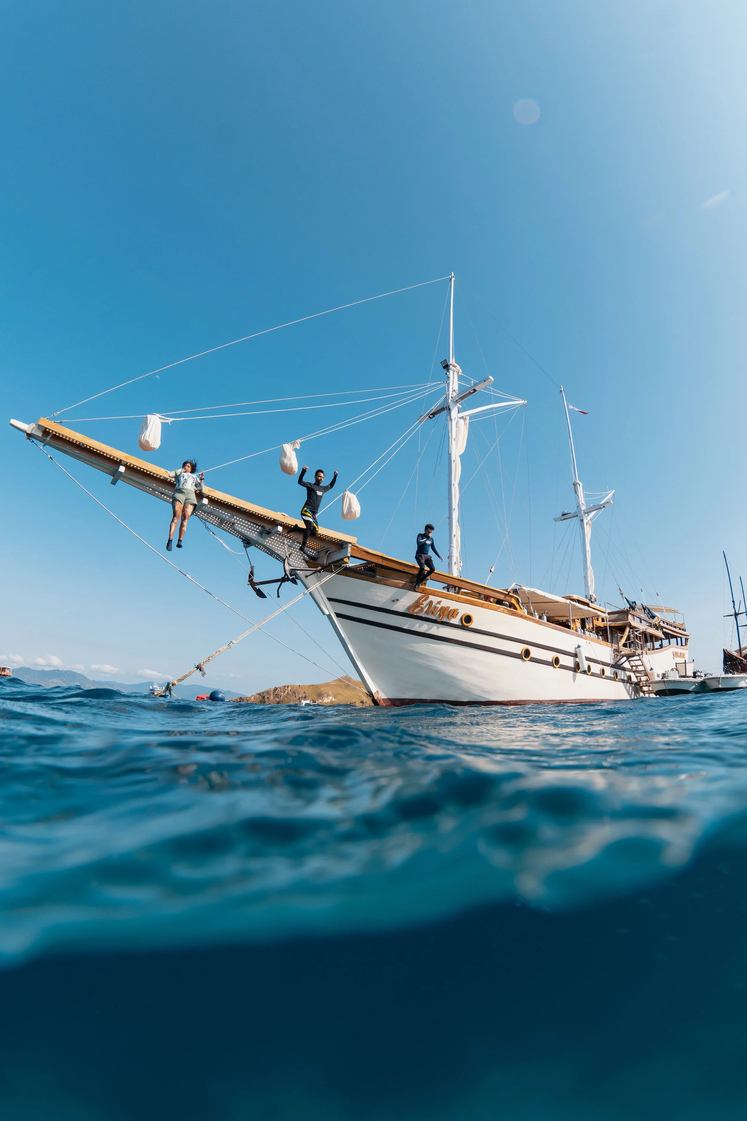 People standing on the bow of a large white sailing ship anchored in the water with a rocky island in the background under a clear blue sky.