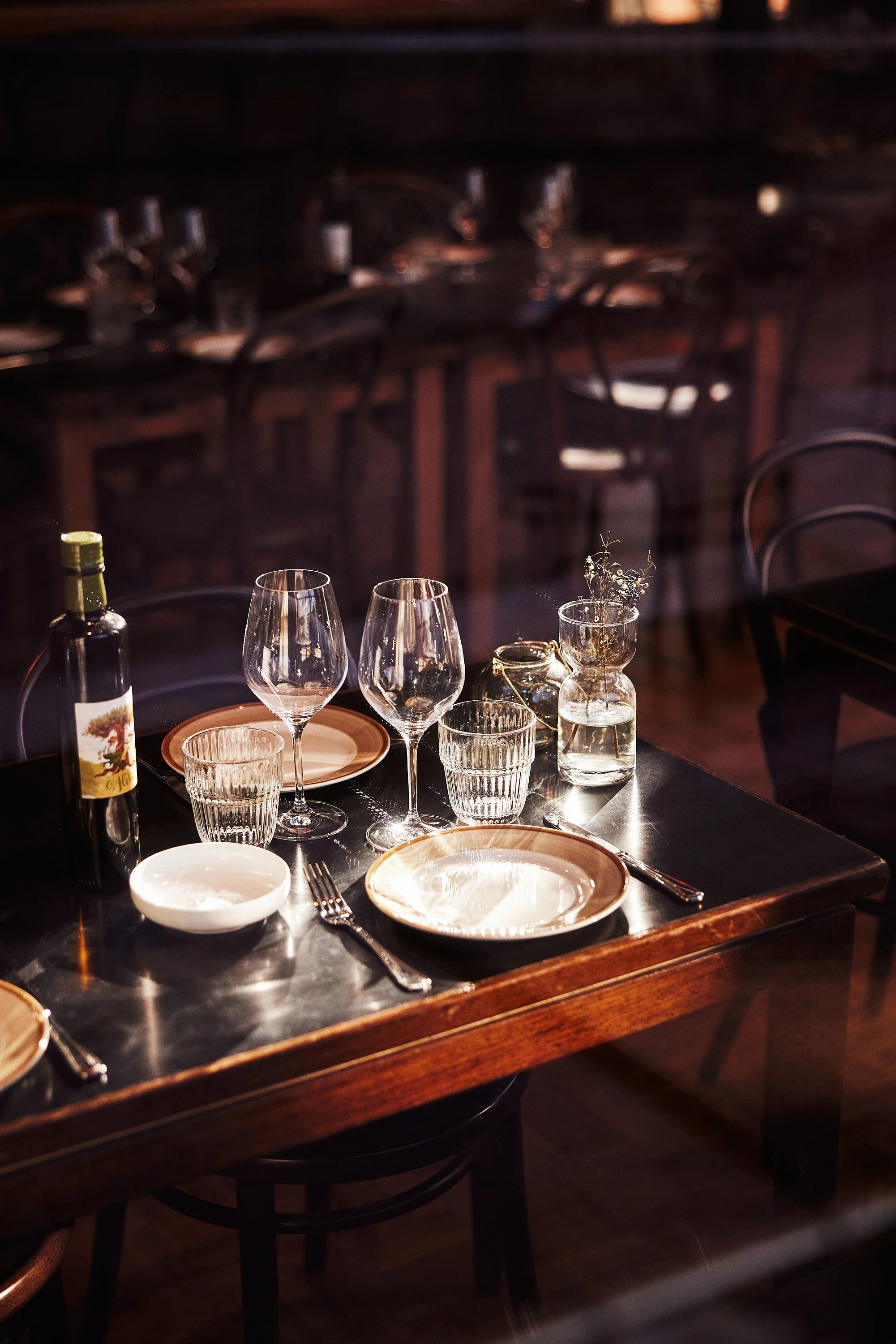 Festbord med glass, tallerkener, bestikk, vaser og vinflaske på et bord i en restaurant, med flere tomme stol rundt.