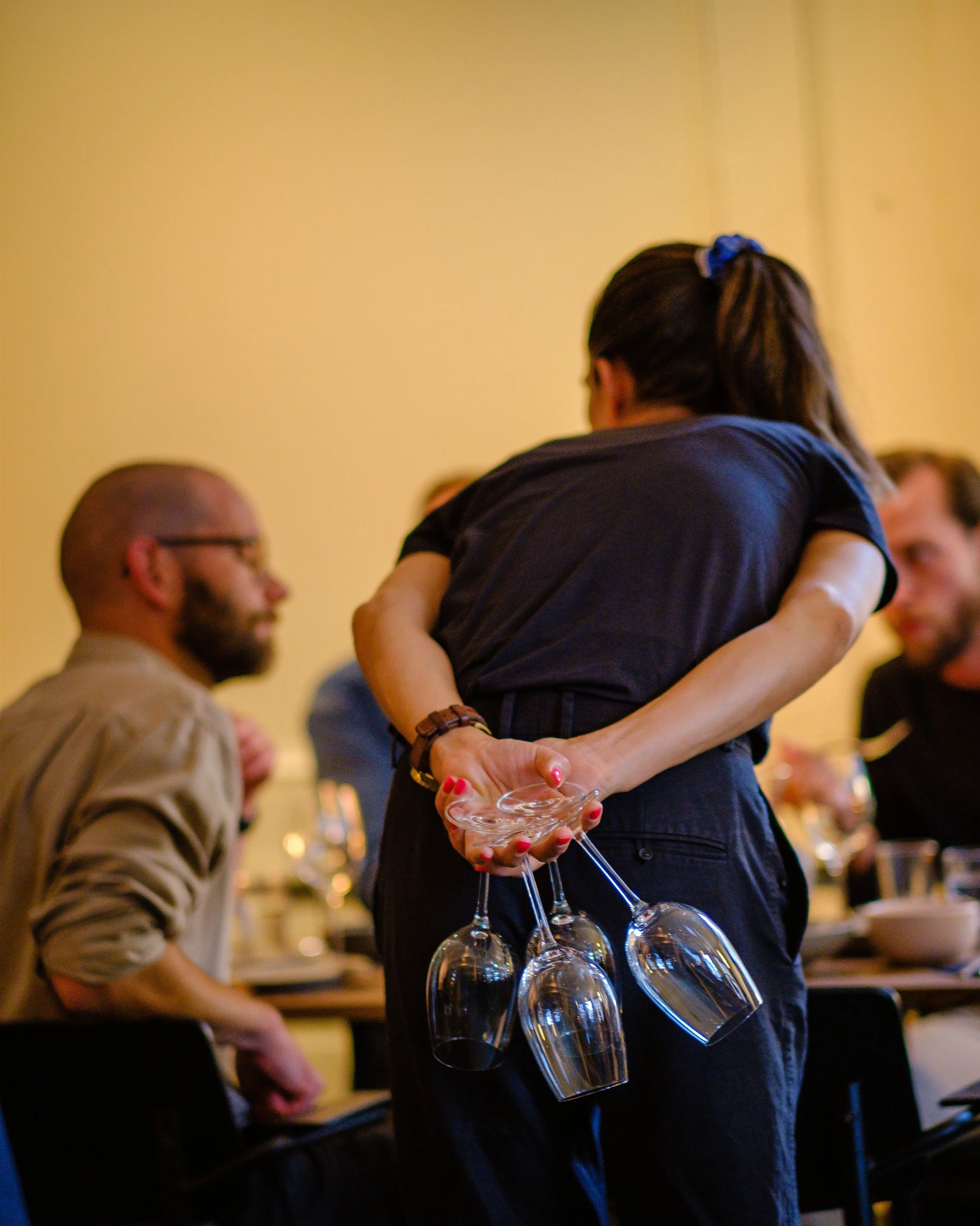 En kvinne som holder et cocktailglass i en bar eller et restaurantmiljø, med flere tomme vinglass hengt opp på beltet hennes, mens andre personer ser ut til å sitte rundt et bord i bakgrunnen.