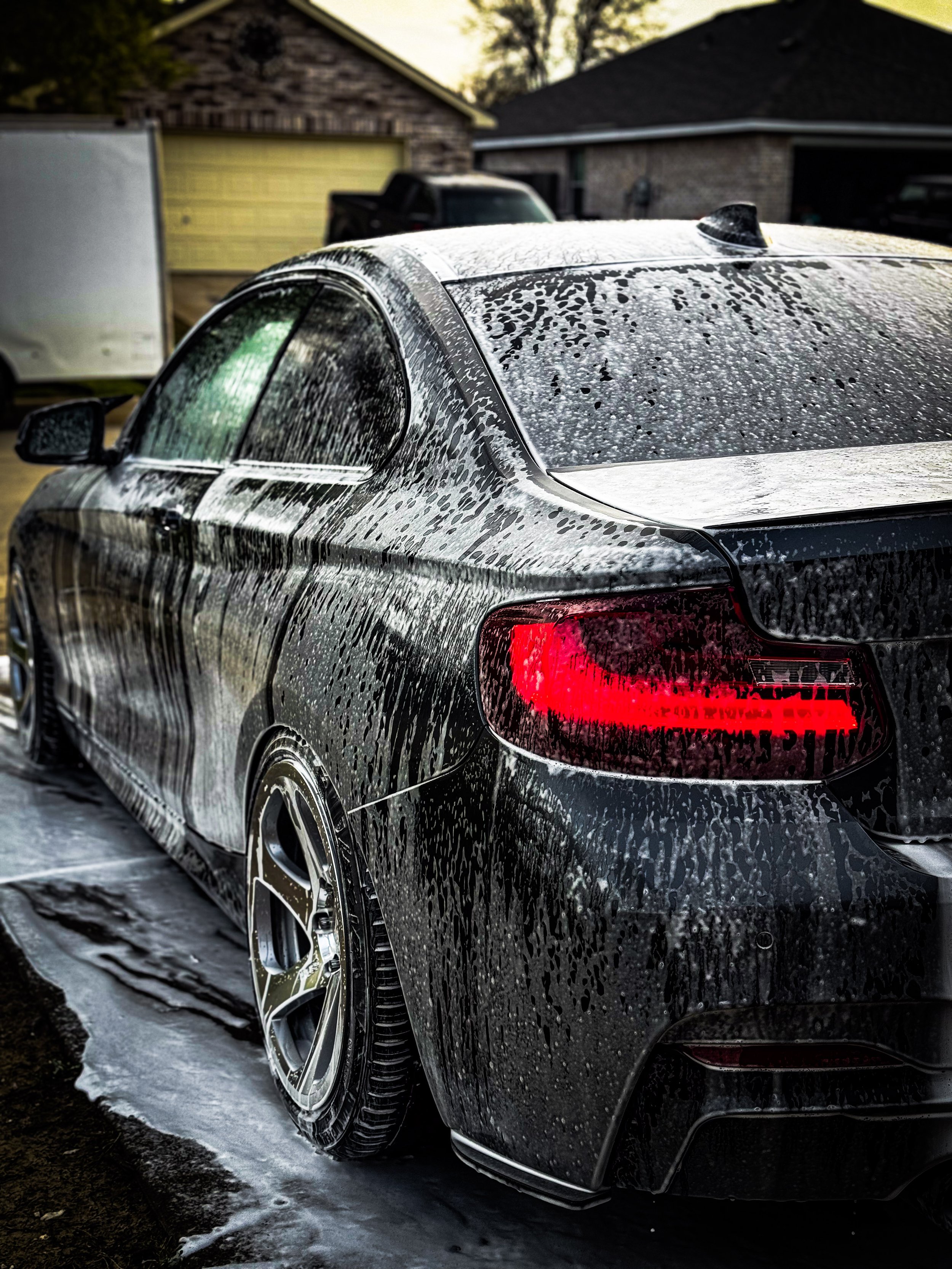 Sleek black car being washed with soapy water, parked in a driveway, red tail light visible. Brick house and garage in the background.