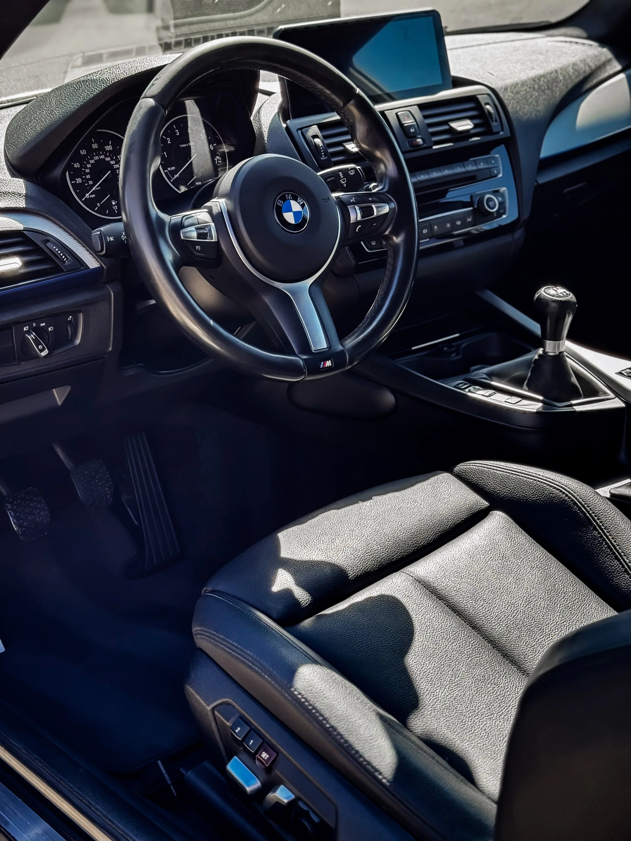 Interior of a BMW car showing the steering wheel, dashboard, manual gear shift, and black leather seat.