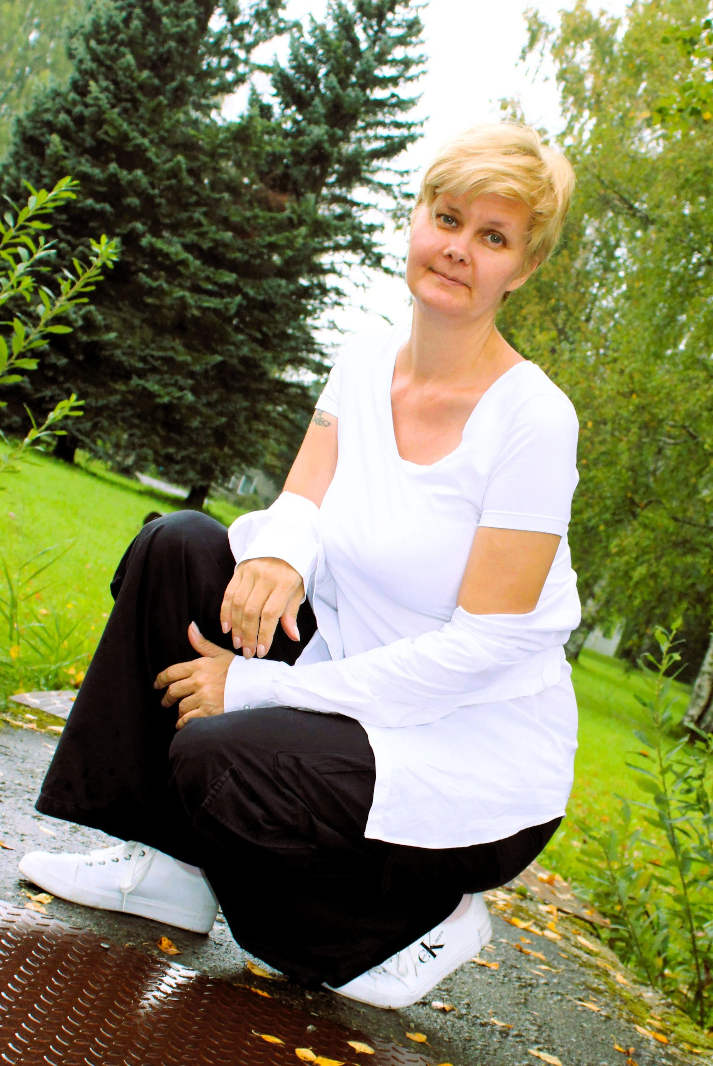 Person with short blonde hair kneeling on a path in a park, wearing a white shirt, black pants, and white sneakers, with green trees and a grassy area in the background.