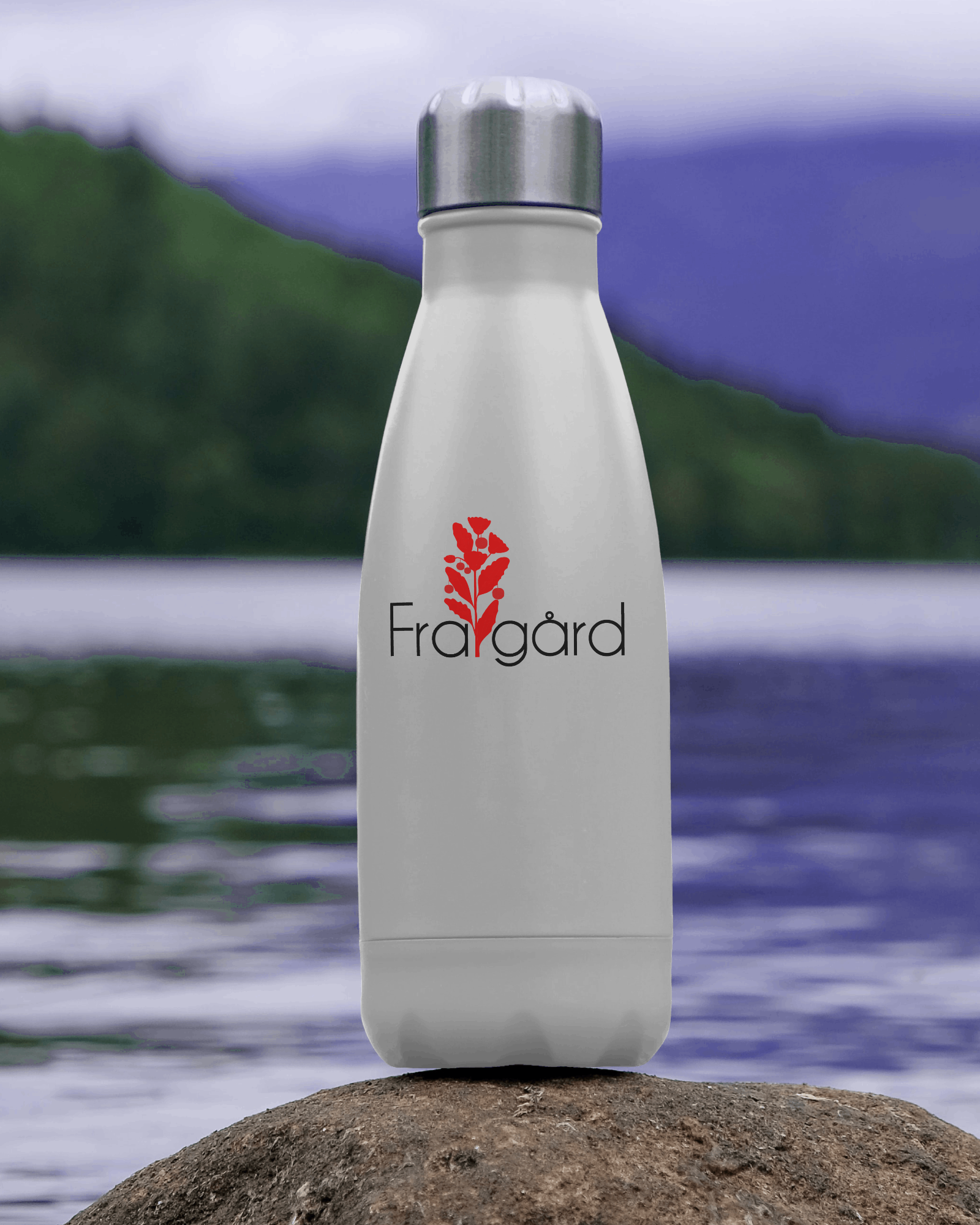 A white water bottle with a silver lid, featuring a red leaf graphic and the black text 'Frailgård' on it, placed on a rock near a body of water with a mountain and cloudy sky in the background.