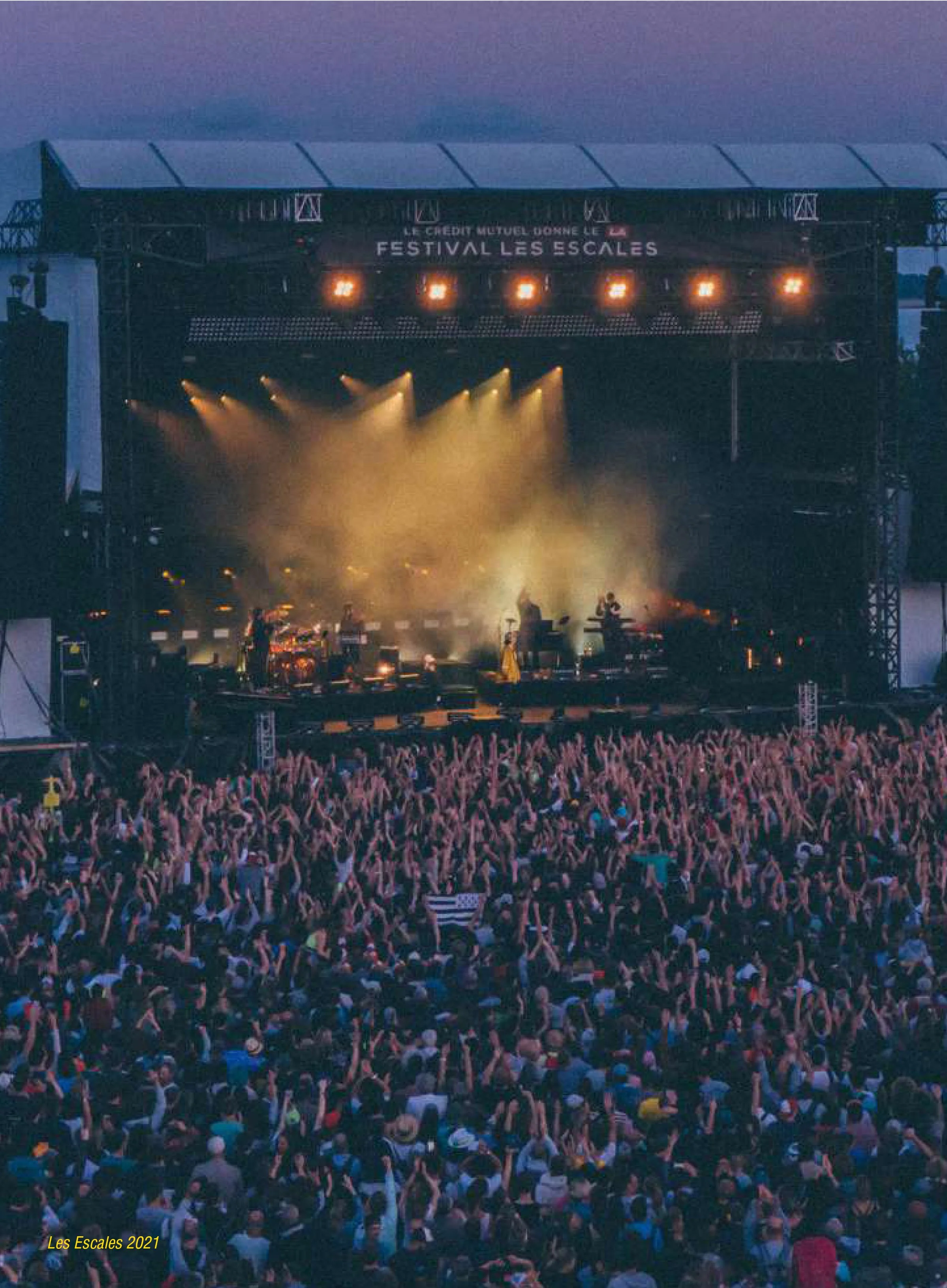 A large crowd gathered at an outdoor concert or festival during evening time, with many people raising their hands. There is a stage with bright orange lights and a banner that reads 'Festival Les Escales' under a clear sky transitioning from day to 