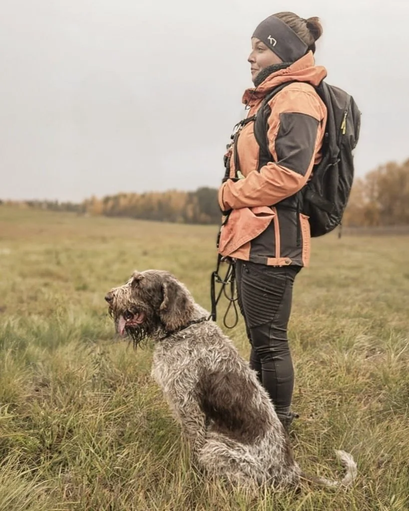 A woman dressed in outdoor gear standing with a leash attached to a large, scruffy dog sitting on a grassy field during daytime.