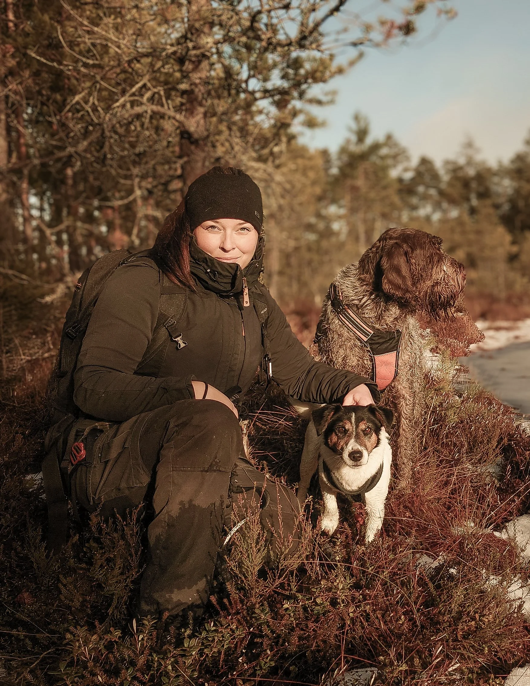 A woman in outdoor winter clothing with two dogs on a wooded lakeshore during sunset.