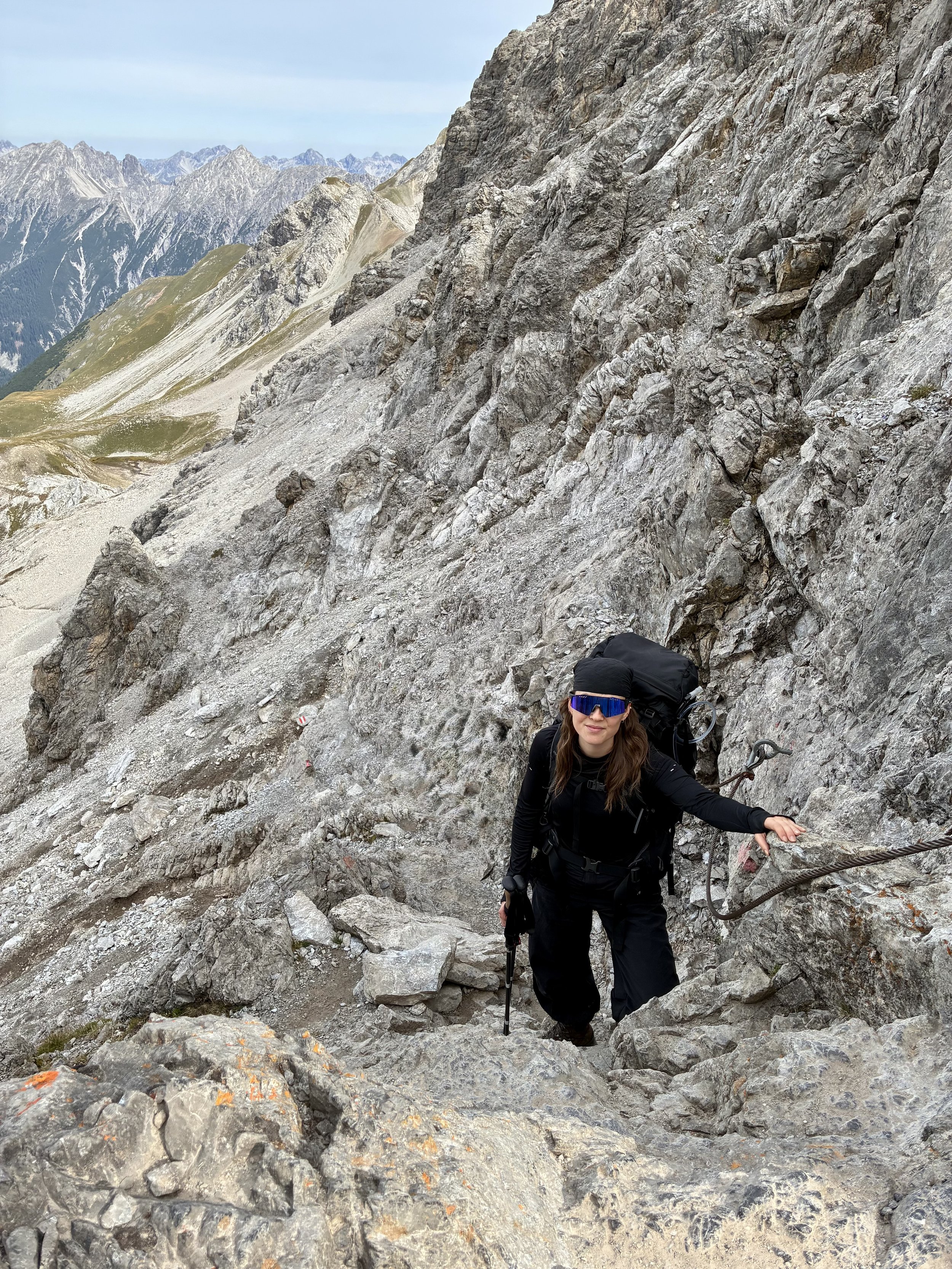 A woman dressed in black hiking gear, wearing sunglasses and a head covering, ascending a steep, rocky mountain trail with safety chains on the rocky wall, with mountain peaks in the background.
