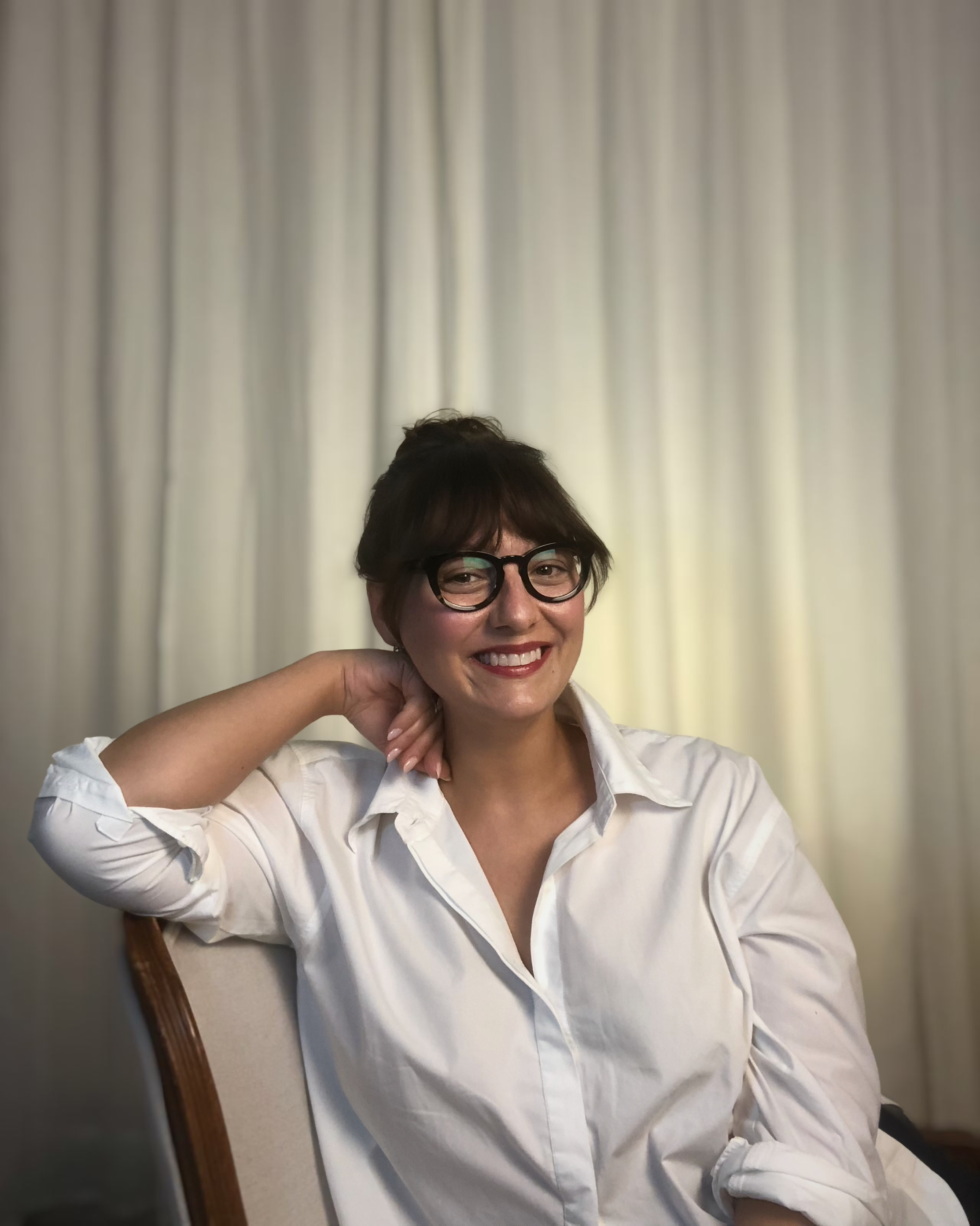 A woman with short brown hair, wearing glasses and a white button-up shirt, smiling while sitting on a chair with her left arm resting on the back. Behind her is a cream-colored curtain.