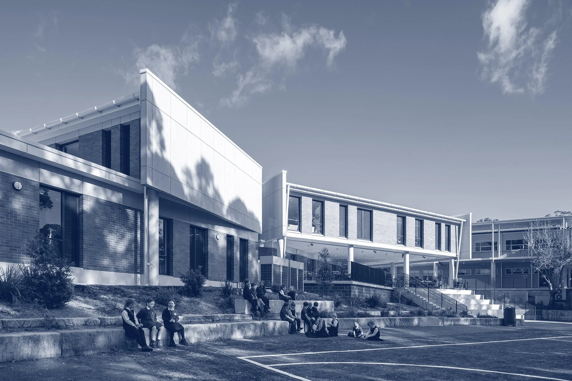 A modern multi-story building with large windows, brick and white panel exterior, and a sloped roof, with children sitting on steps in front of it, under a partly cloudy sky.