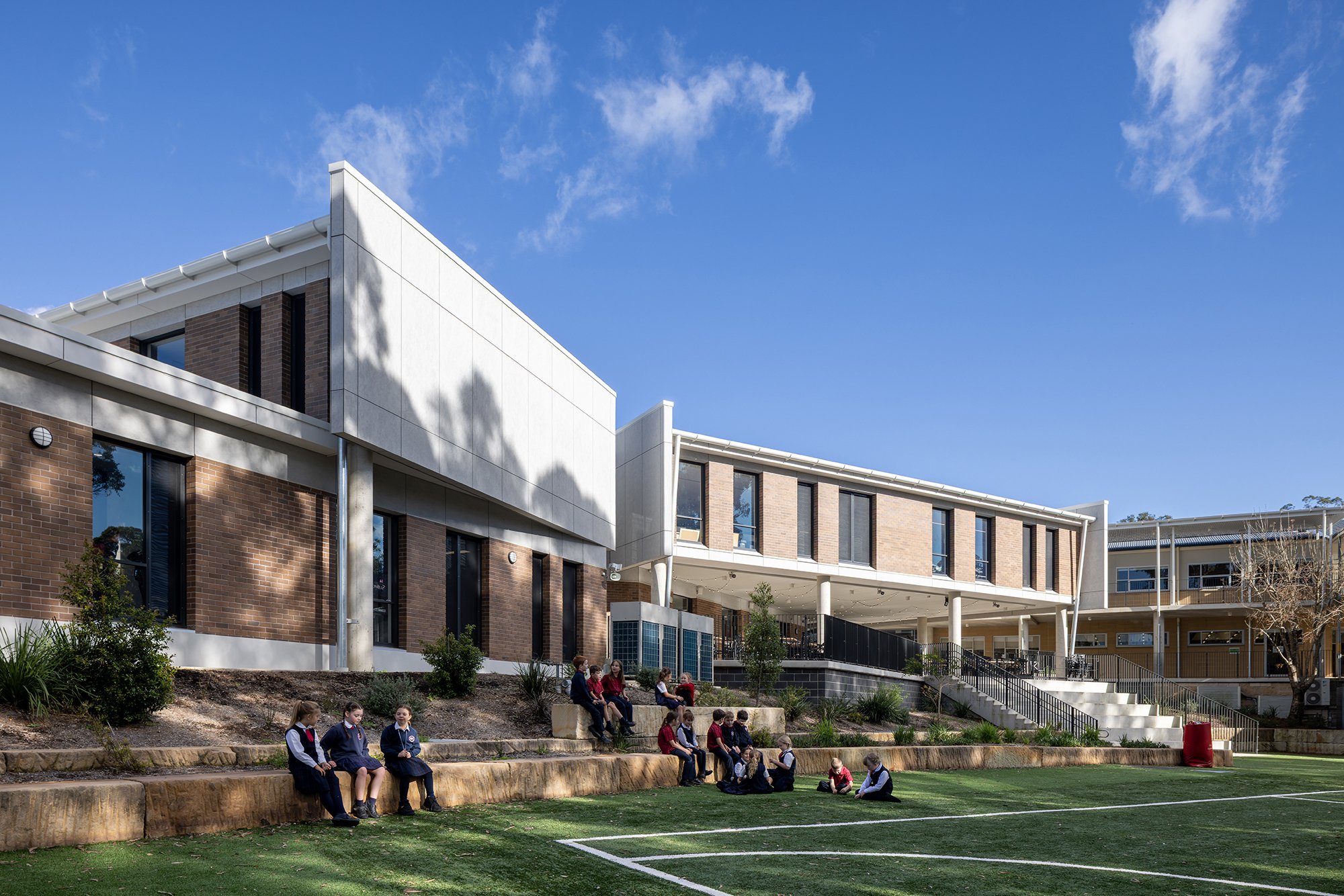 Students sitting on concrete steps and grass outside a modern school building with brick and white exterior walls, large windows, and trees under a partly cloudy blue sky.