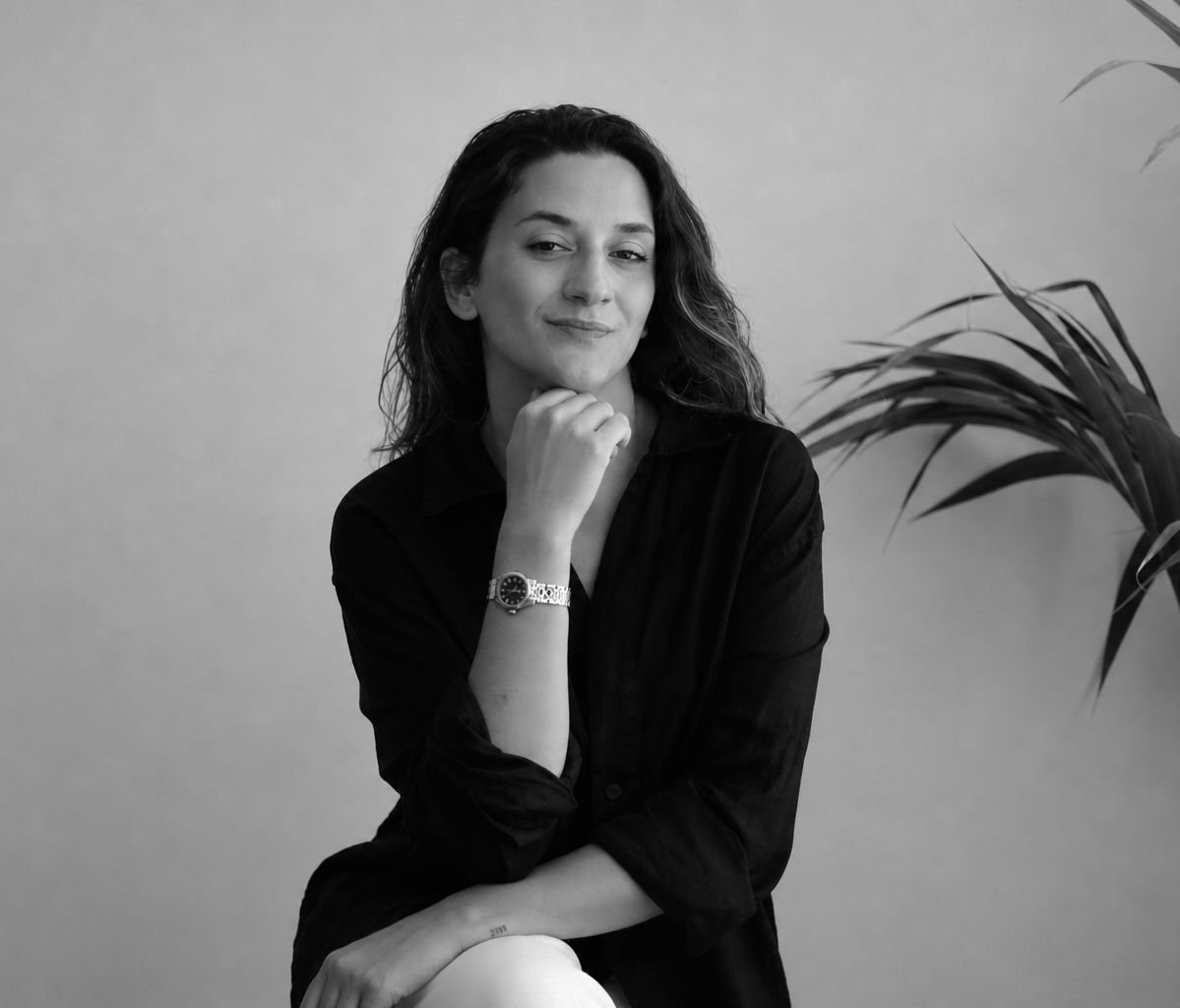 Retrato de una mujer con cabello rizado, vistiendo blusa negra, posando con la mano en el mentón, en un fondo liso con una planta en el lado derecho