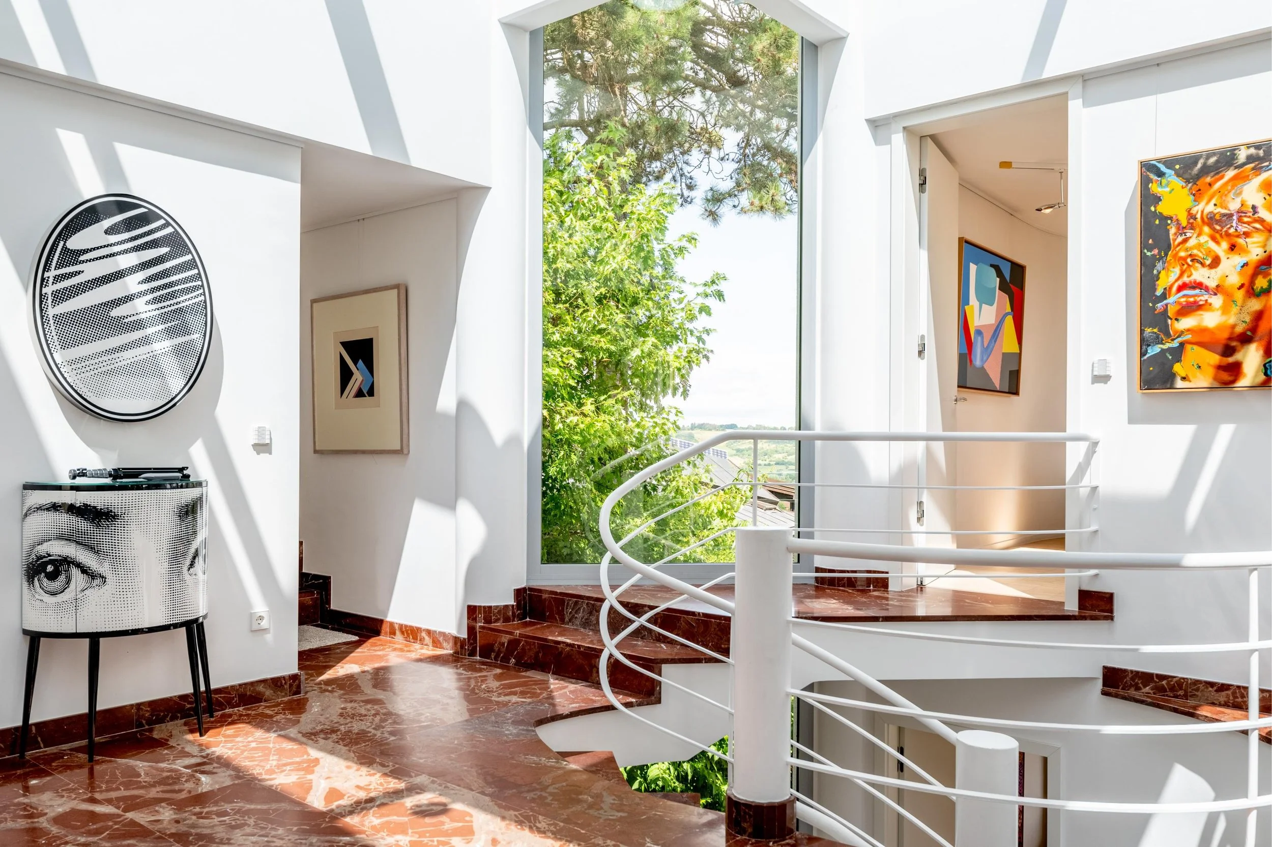 Salle d'exposition avec œuvres d'art modernes, murs blancs, grandes fenêtres laissant entrer la lumière naturelle, sol en marbre rouge et rampes blanches en spirale.