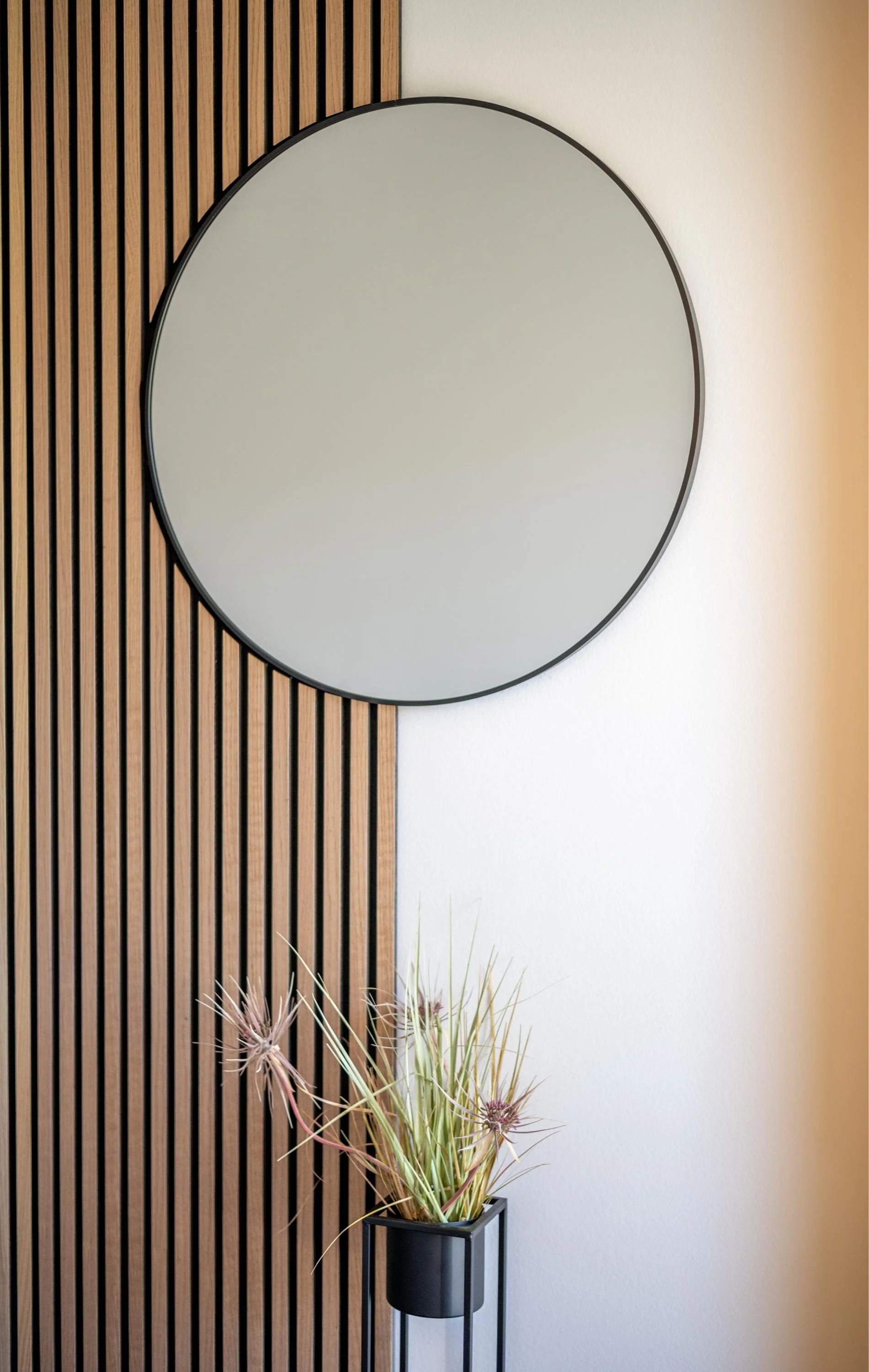 Un miroir rond avec un cadre noir suspendu sur un mur blanc, avec un panneau en bois à rayures verticales à gauche. En dessous, une plante sèche dans un vase noir sur un socle métallique.