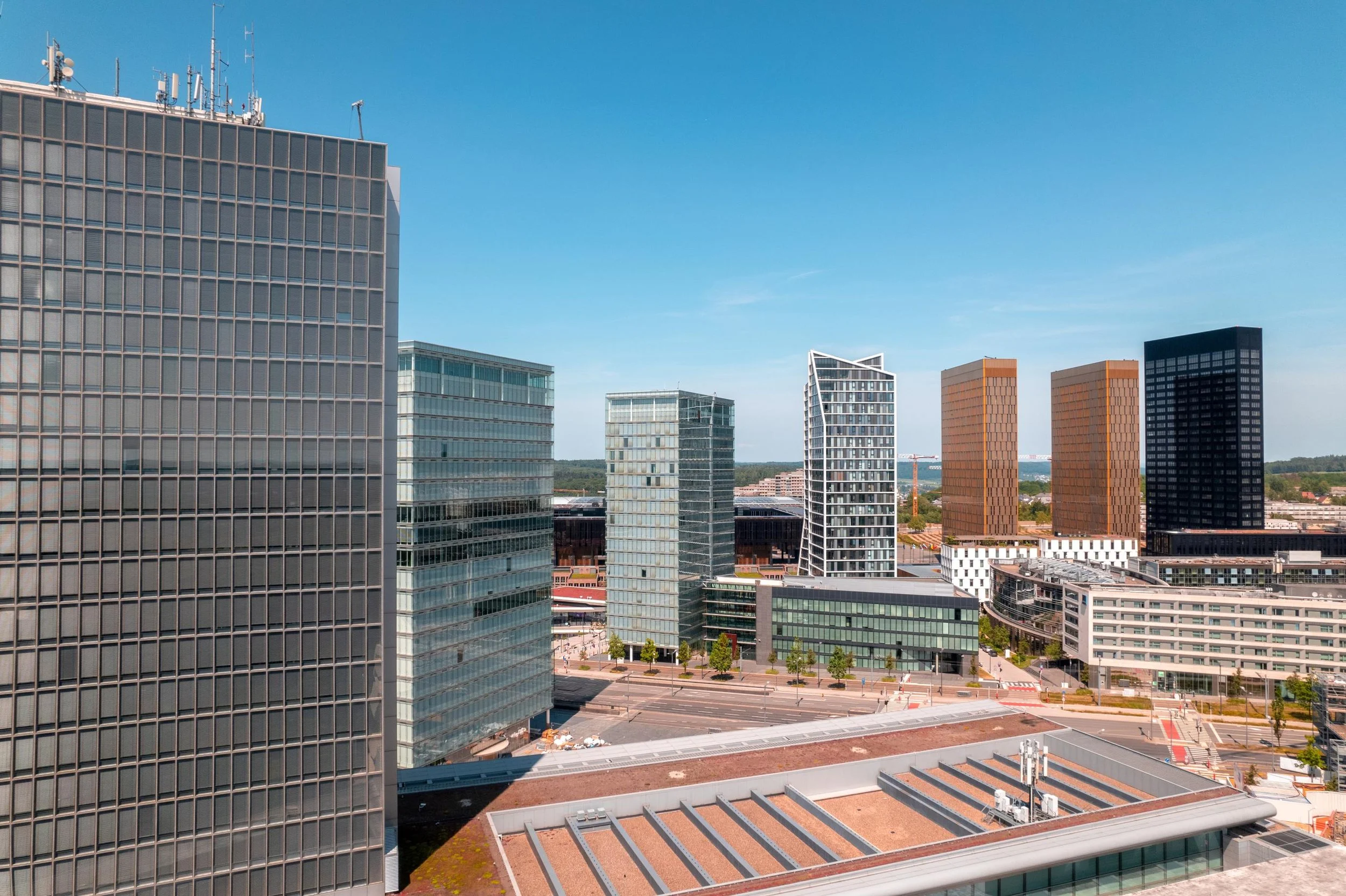 Vue aérienne d'un quartier d'affaires avec plusieurs gratte-ciels modernes en verre, une rue large en dessous et un ciel bleu clair.