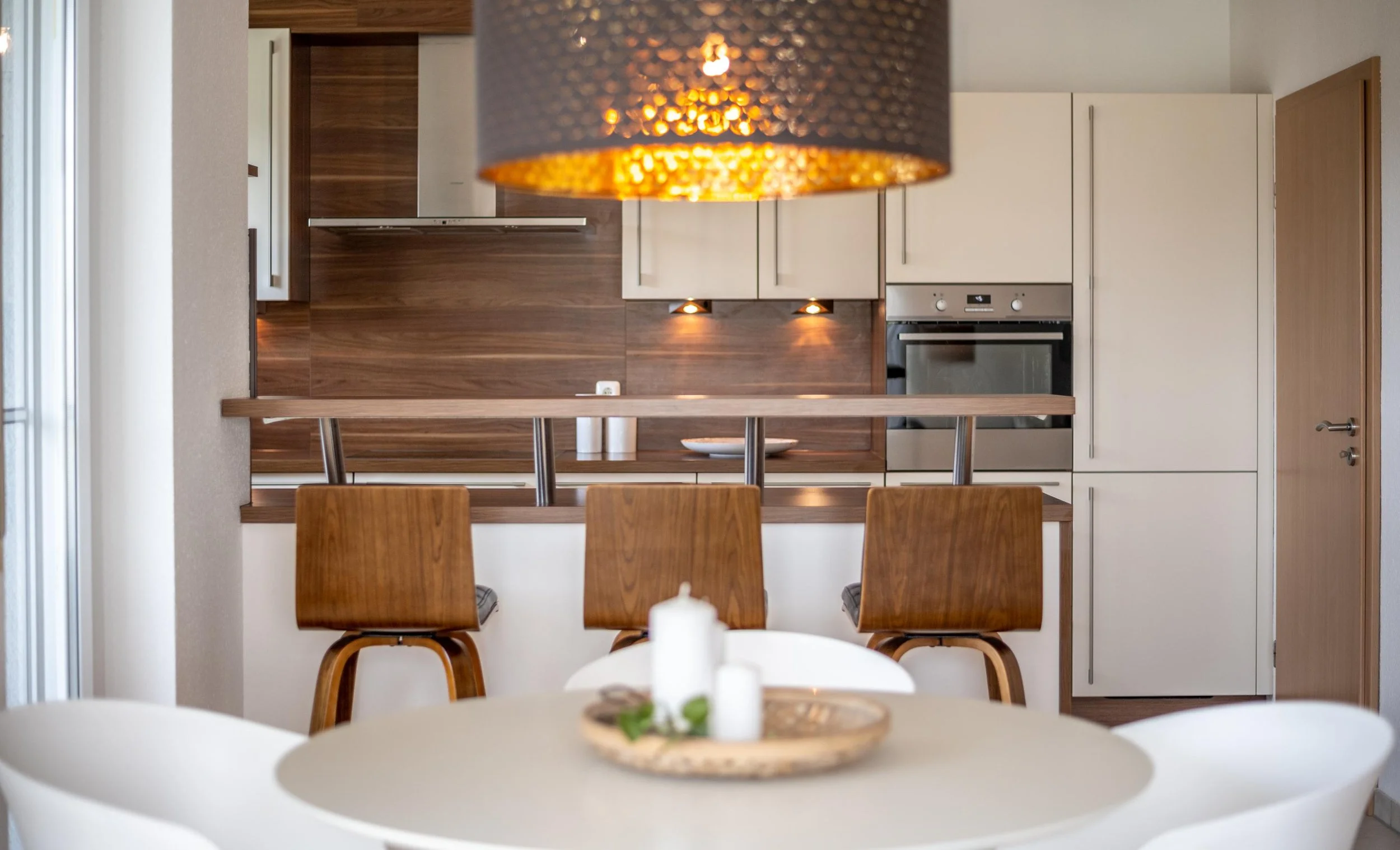 Cuisine moderne avec plan de travail en bois, chaises en bois autour d'une table, luminaire suspendu au plafond, appareils électroménagers intégrés, décor épuré et chaleureux