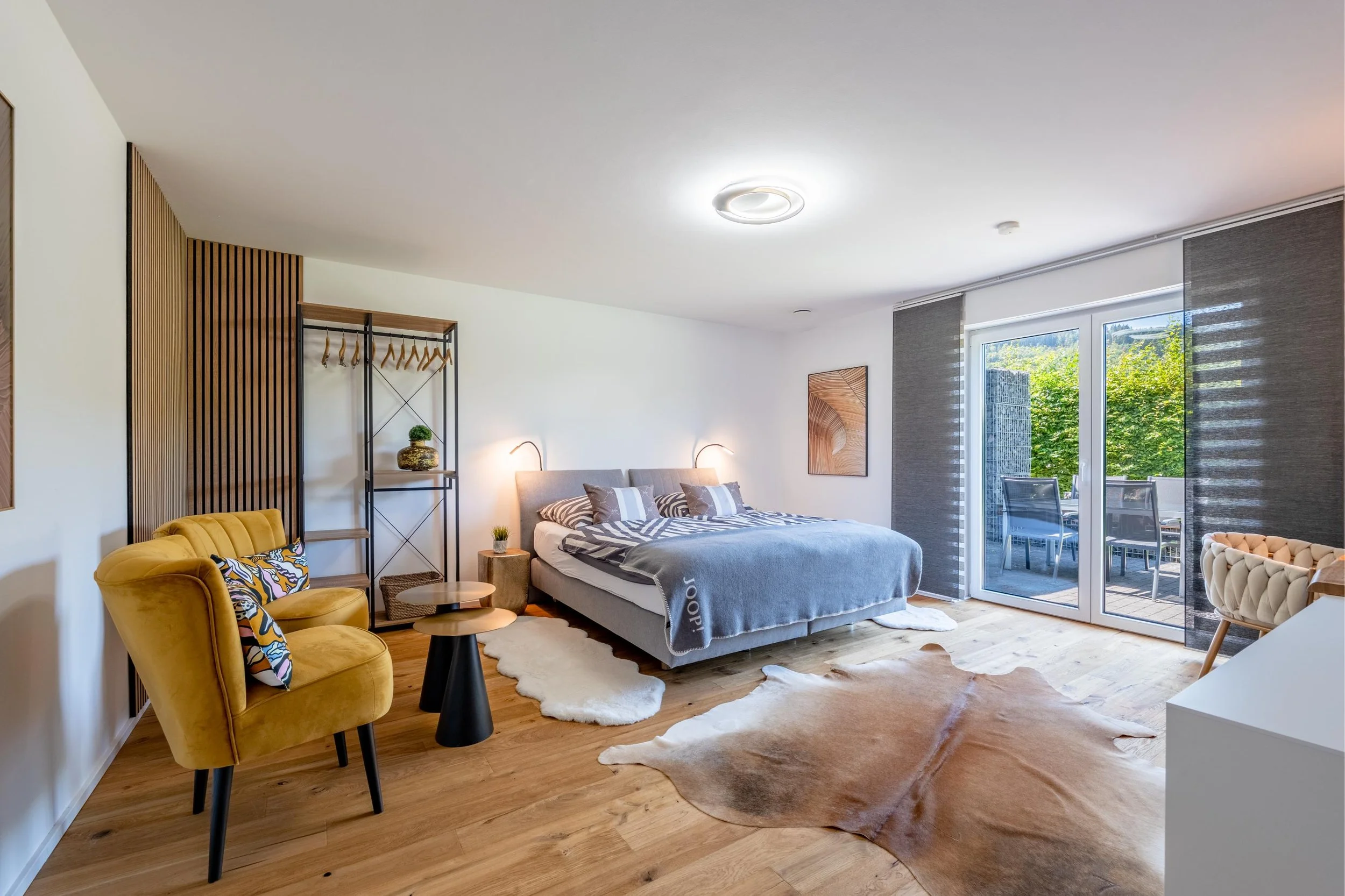 Chambre lumineuse avec un lit, fauteuil jaune, décoration moderne et grande fenêtre donnant sur un balcon extérieur.