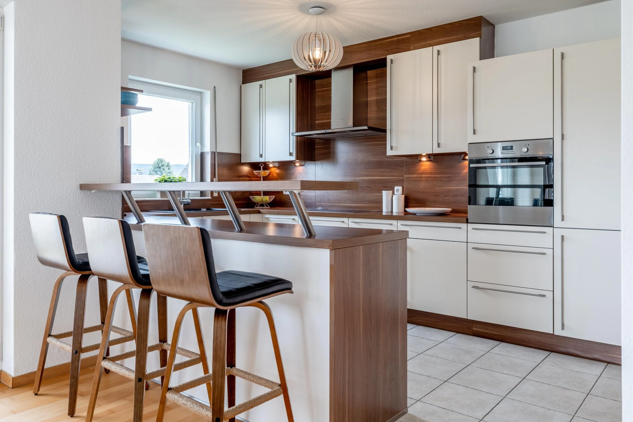 Cuisine moderne avec îlot en bois, chaises de bar en bois, armoires blanches, mur en bois, four encastré, éclairage intégré et fenêtre présentant un paysage extérieur.